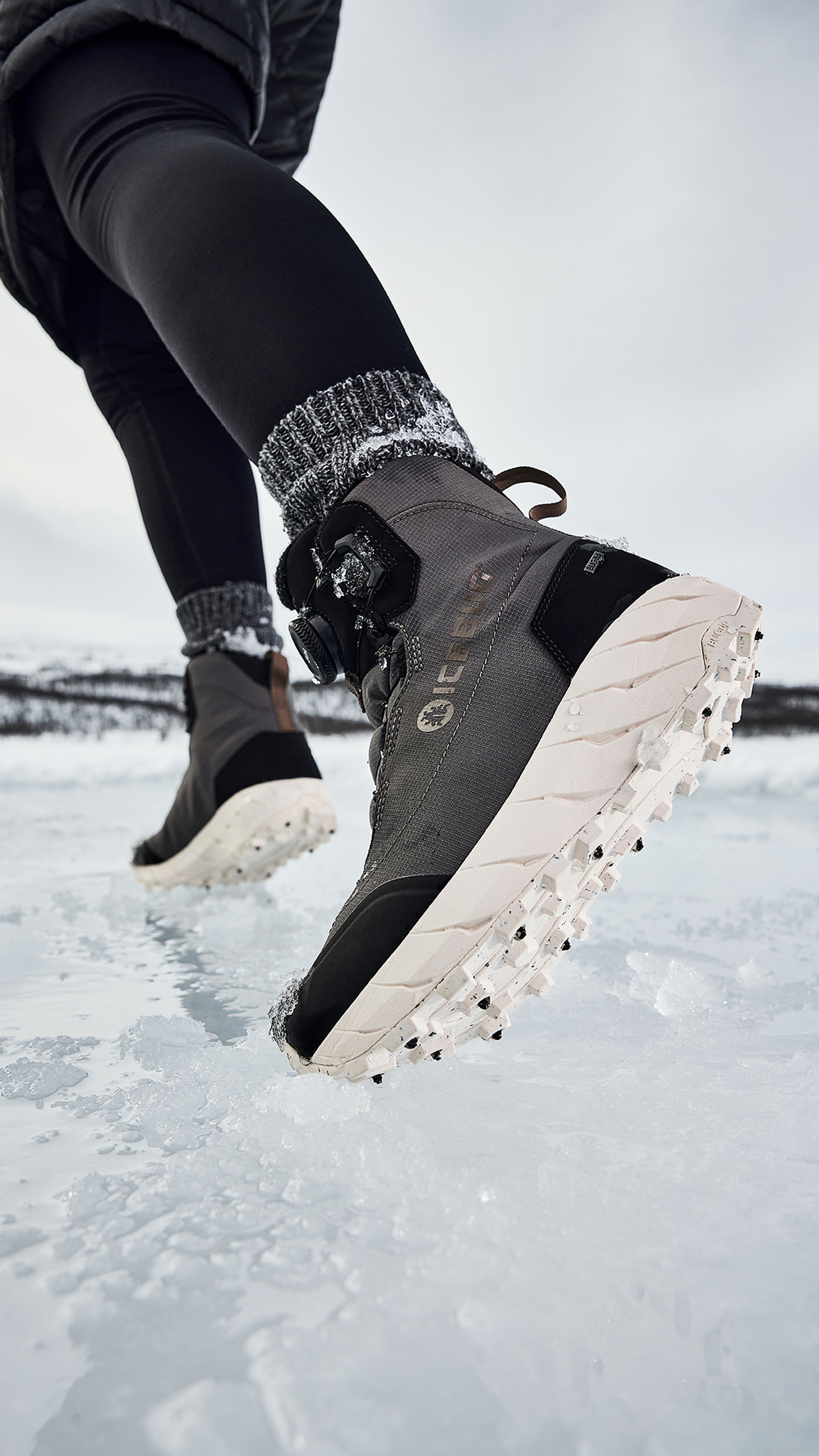 Close-up of a person walking on snow, wearing black leggings and gray winter boots with thick soles, against a cloudy sky background.