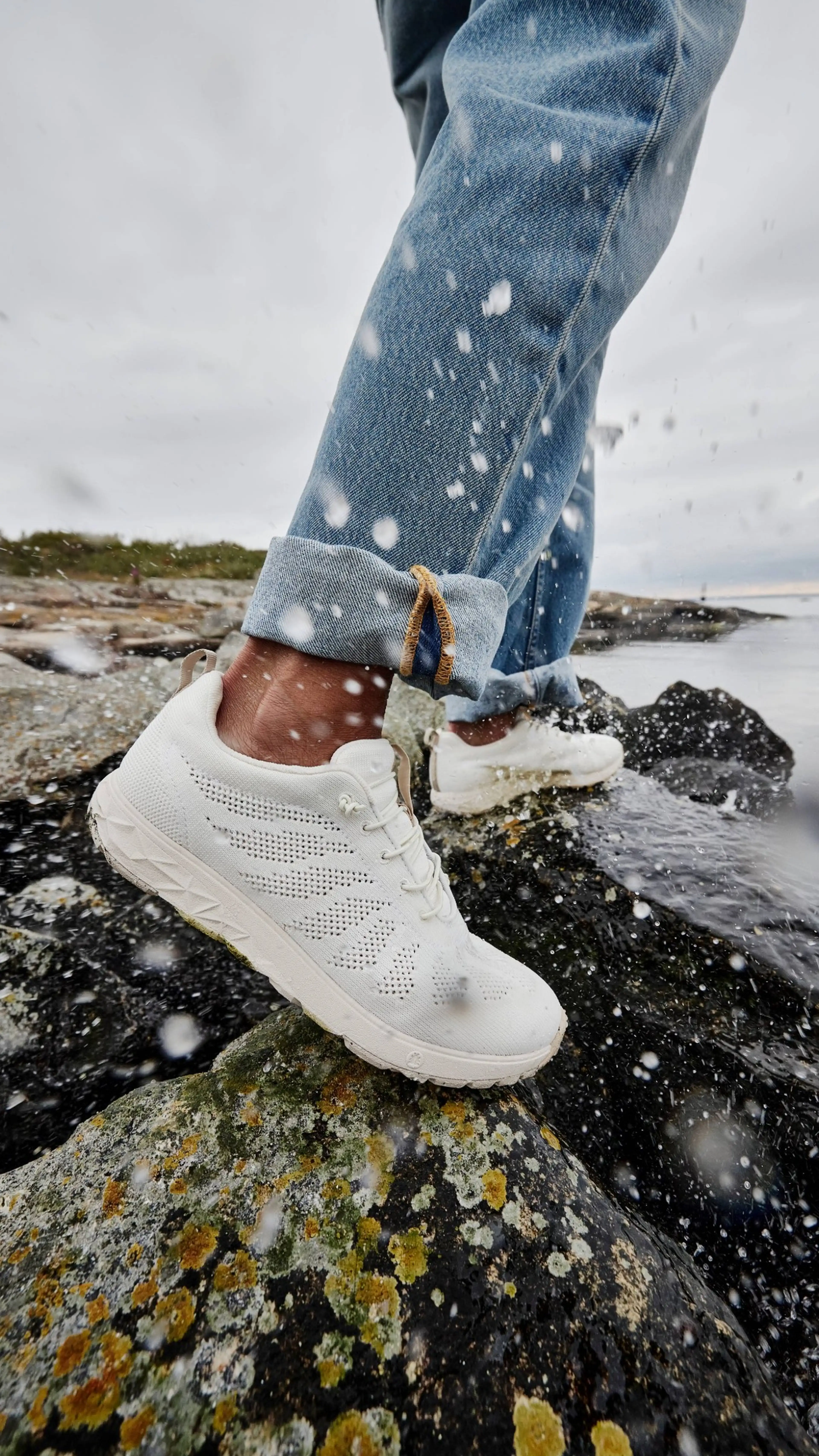 Person in white sneakers and jeans walking on wet, lichen-covered rocks by the sea, with water splashes around.