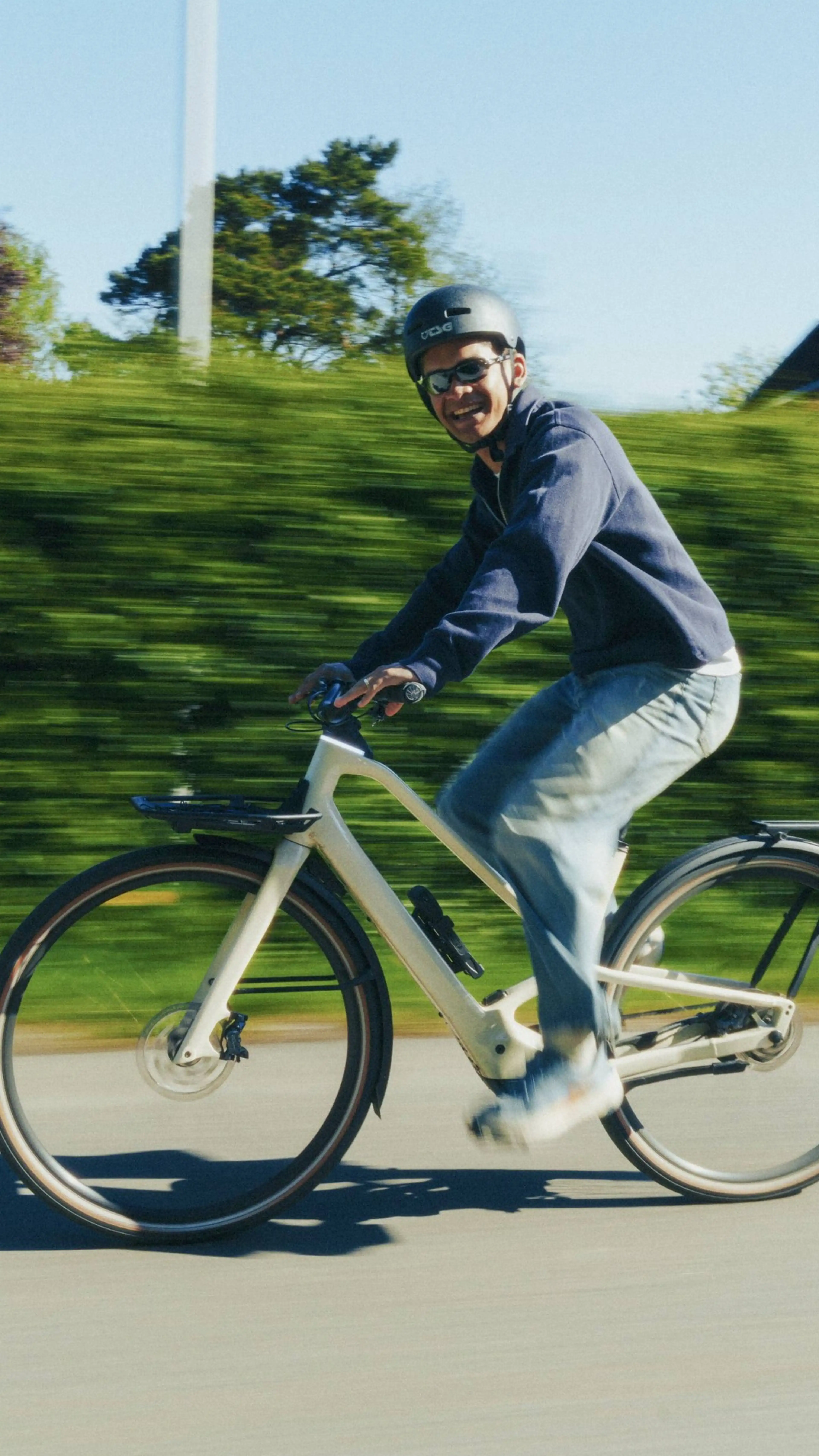 A person in a helmet rides a bicycle at speed on a sunny day, with greenery blurred in the background.