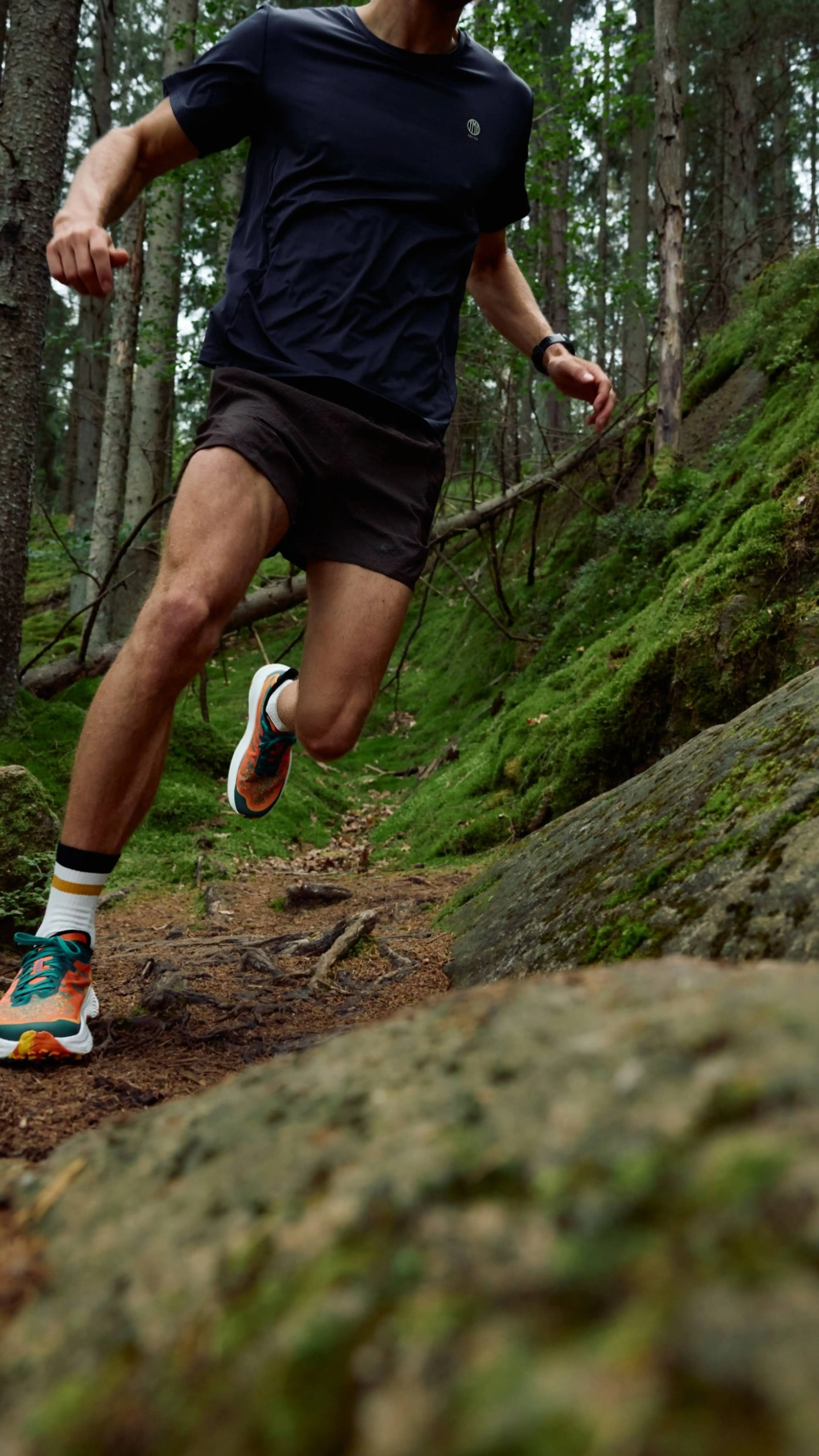 Person trail running in a forest, wearing a dark shirt and shorts with colorful sneakers, surrounded by moss-covered rocks and trees.