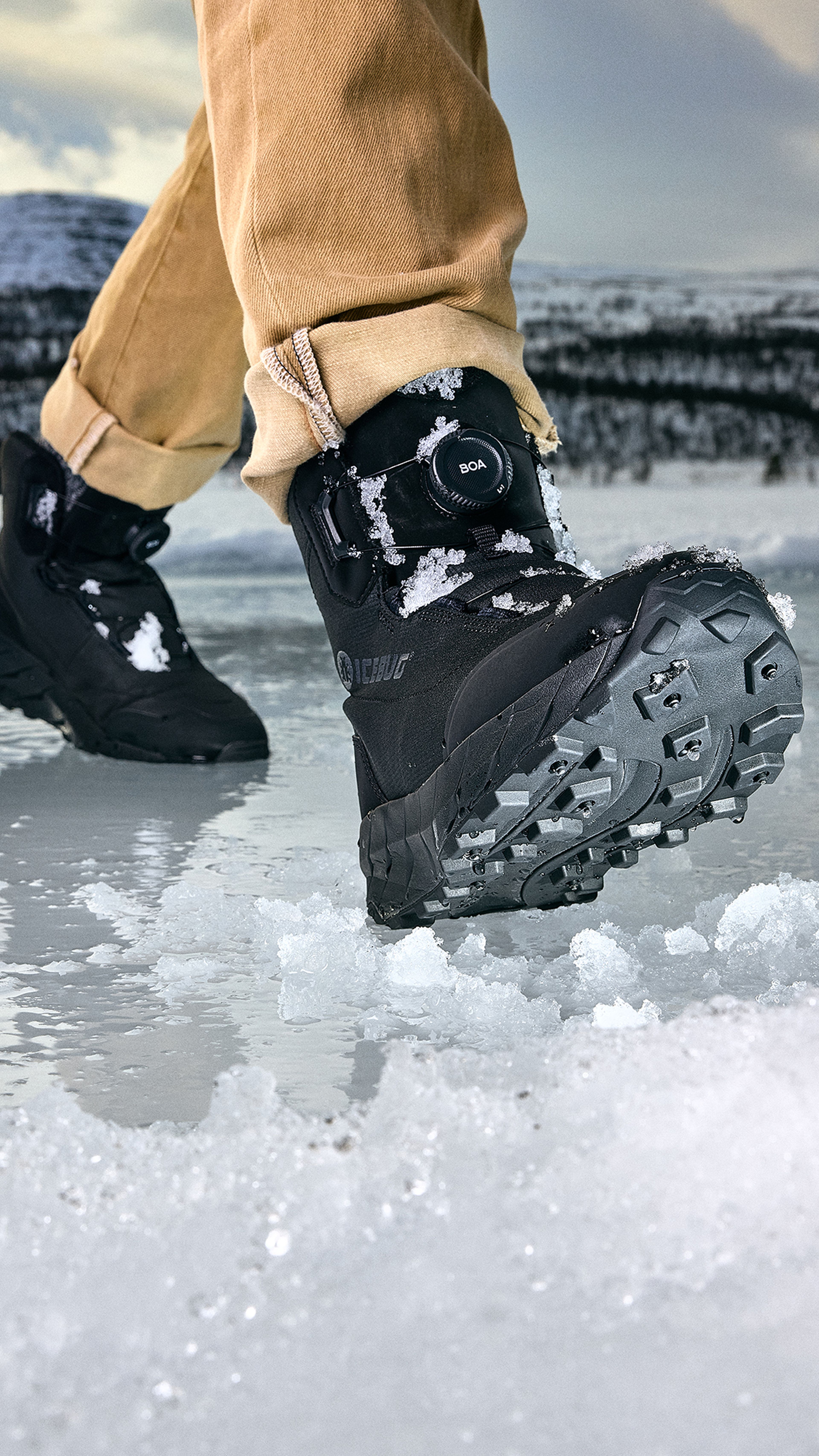 A person wearing tan pants and black winter boots with rugged soles walks on icy ground, with a snowy landscape in the background.