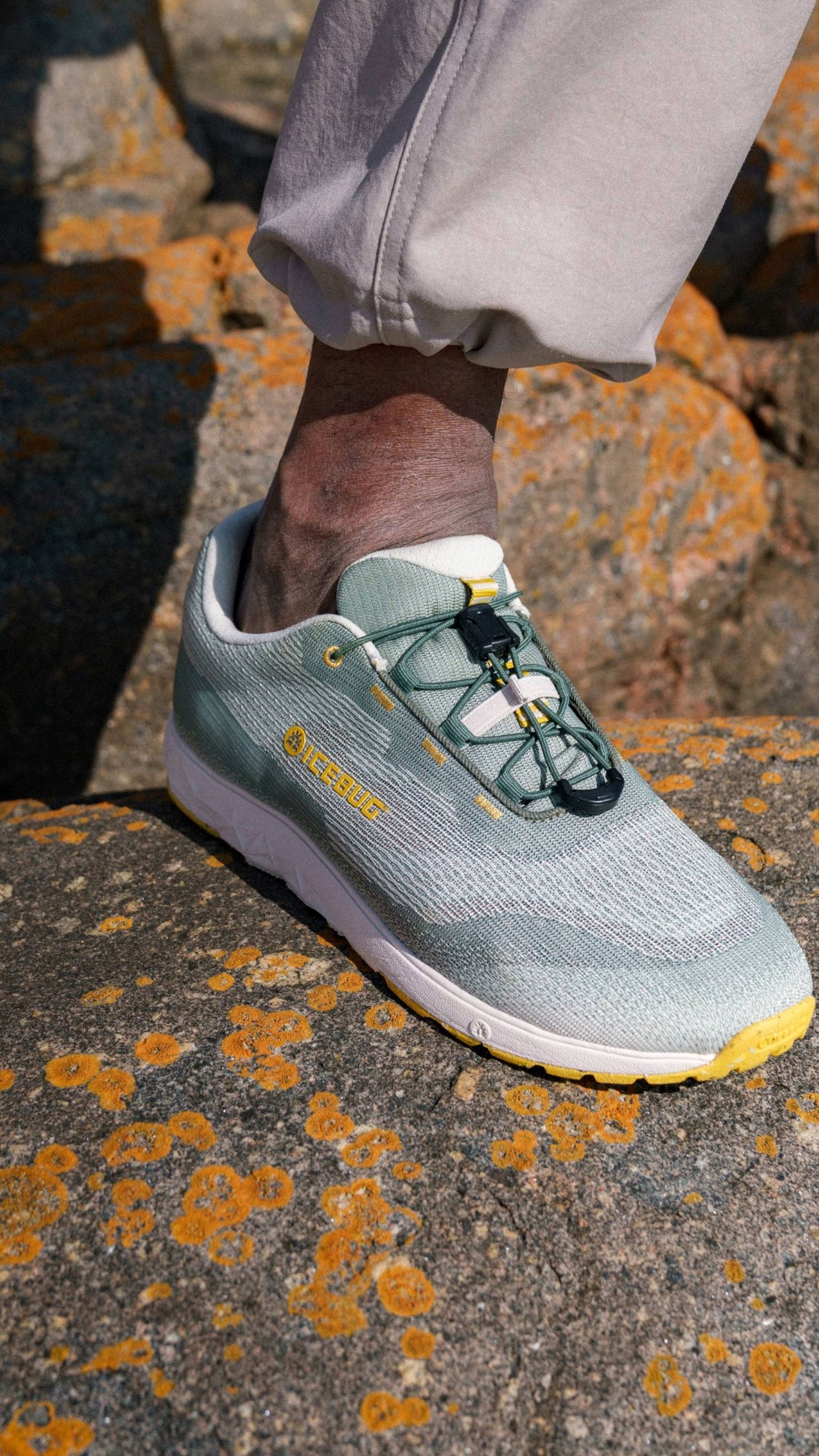 Person wearing light green sneakers with yellow accents, standing on a rocky surface covered in orange lichen.
