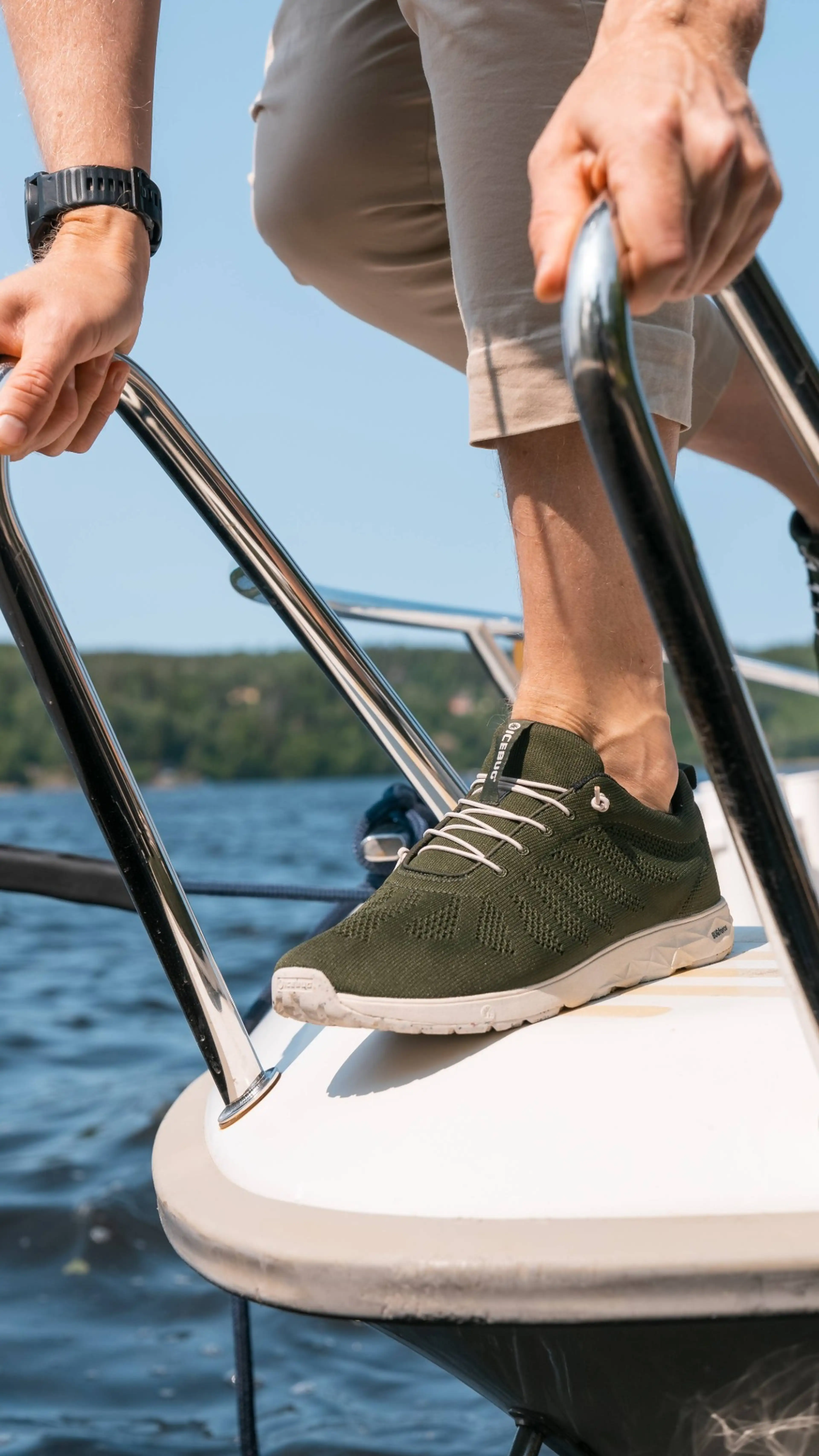Person wearing green sneakers and light shorts stands on the edge of a boat, holding the railing, with a lake and trees in the background.