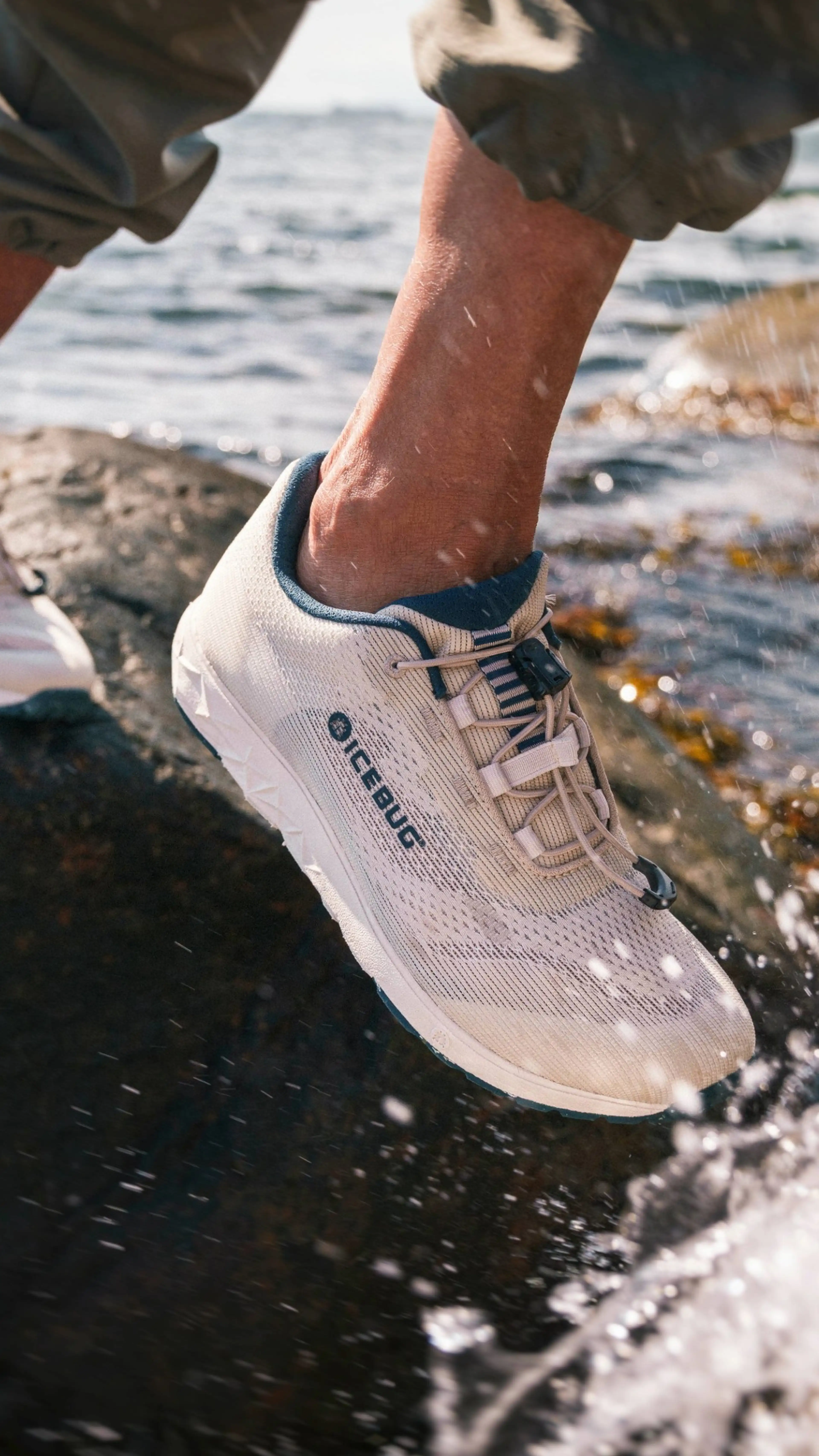 Close-up of a person wearing beige sneakers stepping on a wet rock near the water, with droplets splashing around.