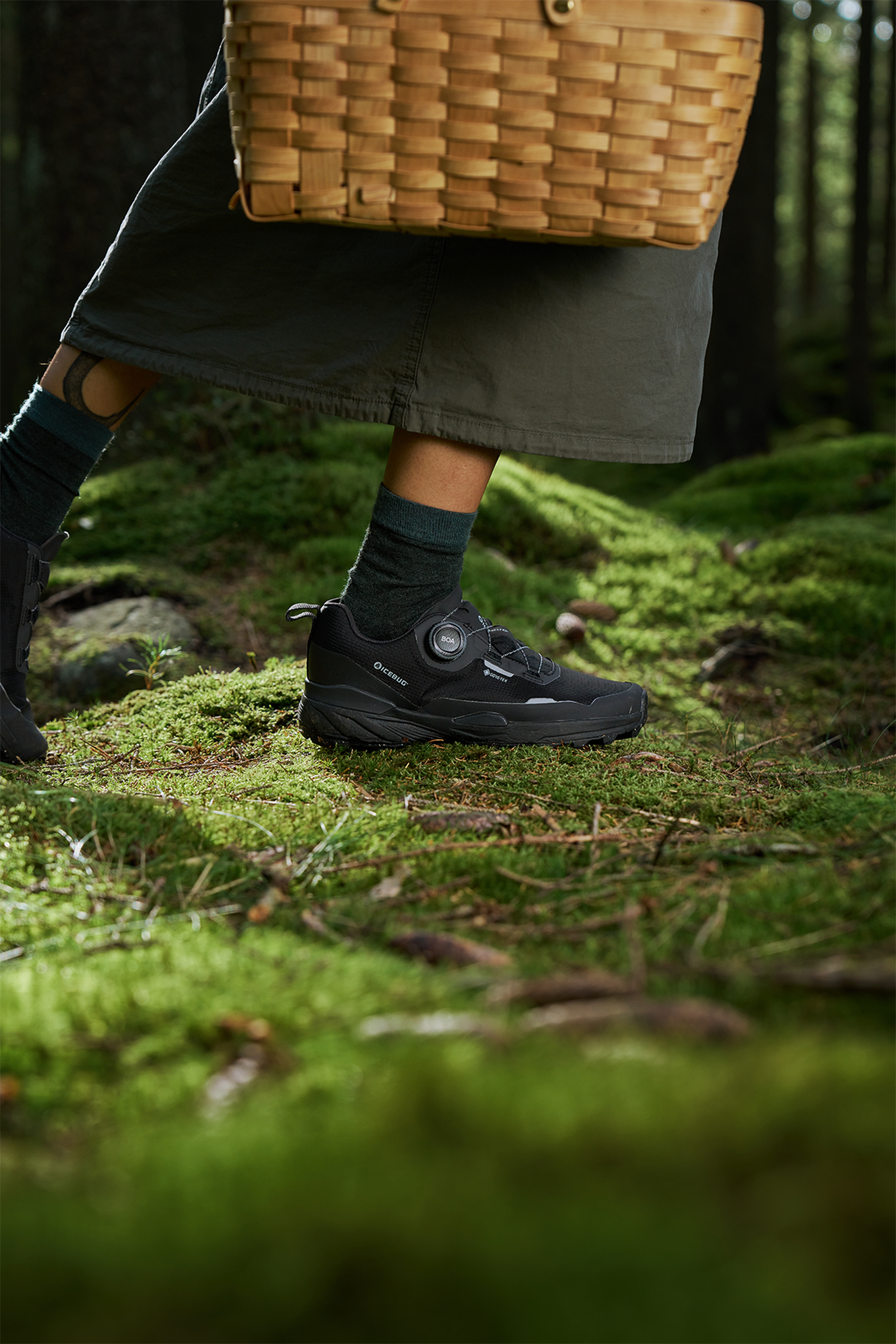 A person in black hiking shoes and green socks walks on a mossy forest floor, carrying a woven basket.