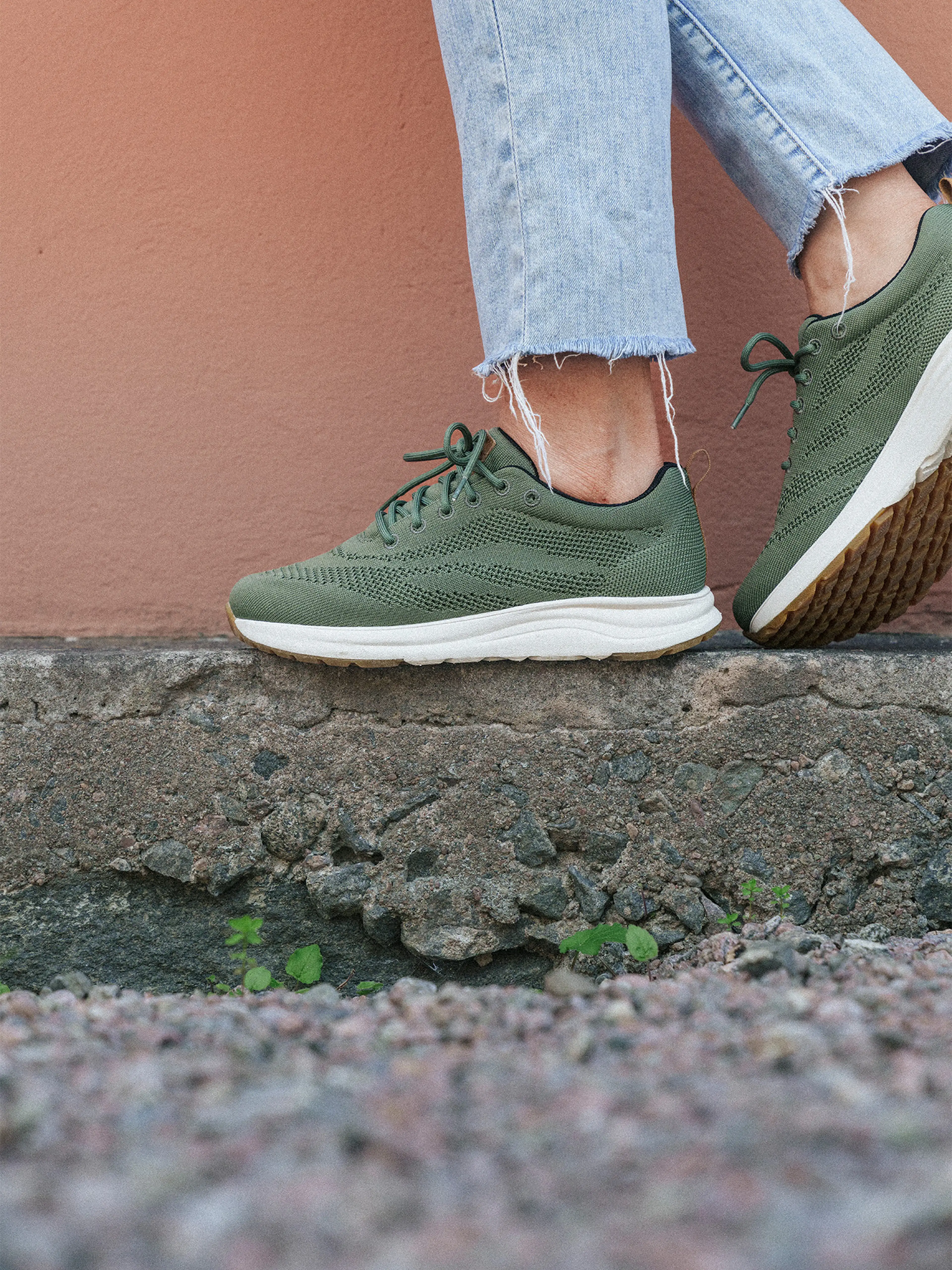 Person wearing green sneakers and frayed jeans standing on a rough stone surface against a peach-colored wall.