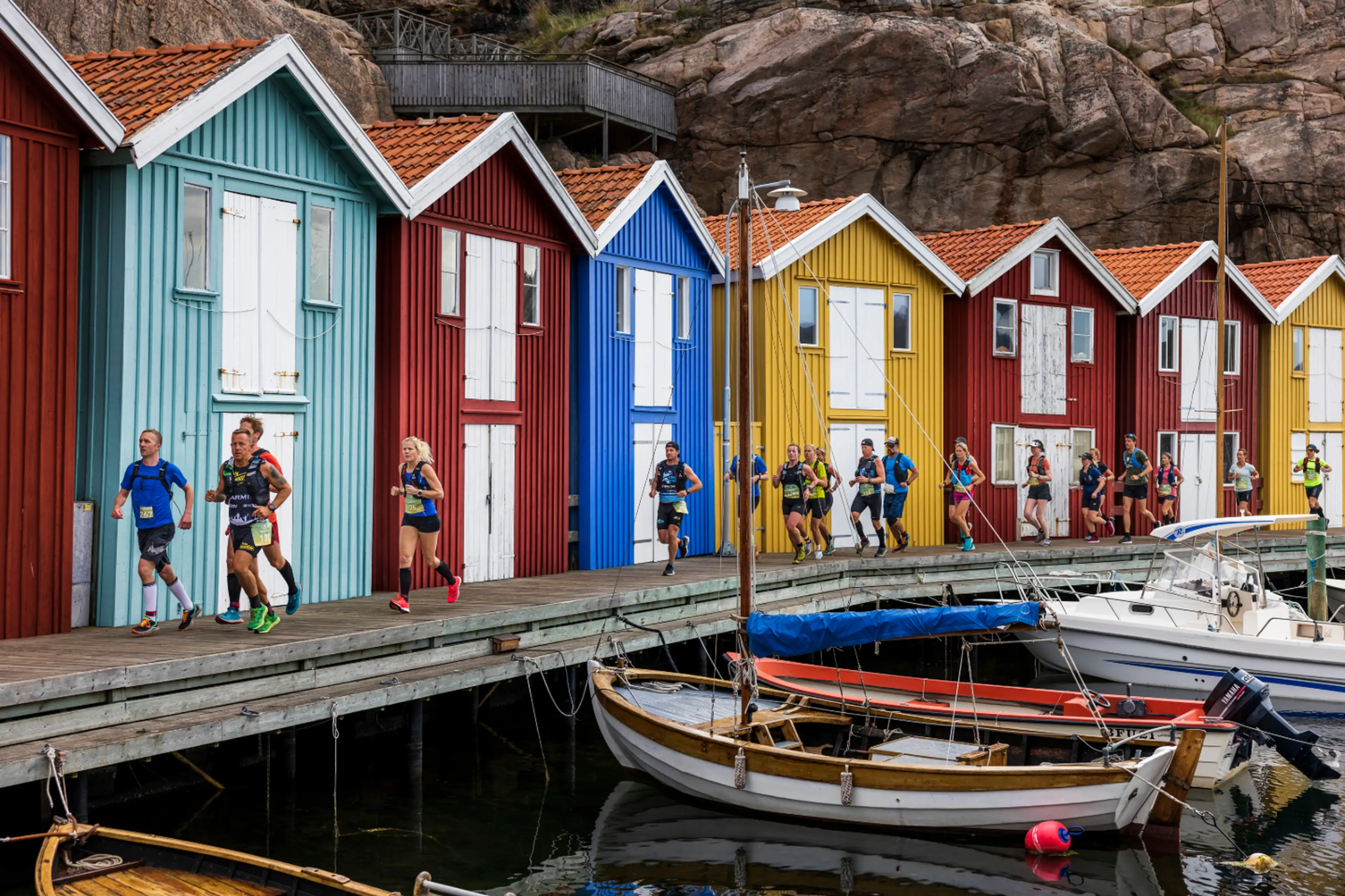 Runners in a race pass colorful wooden houses on a waterfront boardwalk, with boats moored in the foreground and rocky cliffs behind.