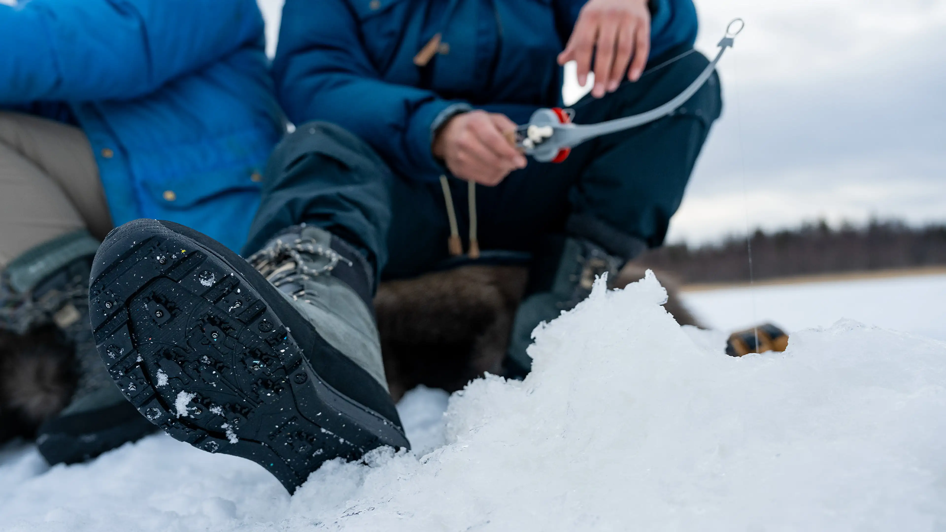 Person ice fishing, sitting on snow with a fishing rod. Close-up of boots and ice hole, cloudy sky in the background.