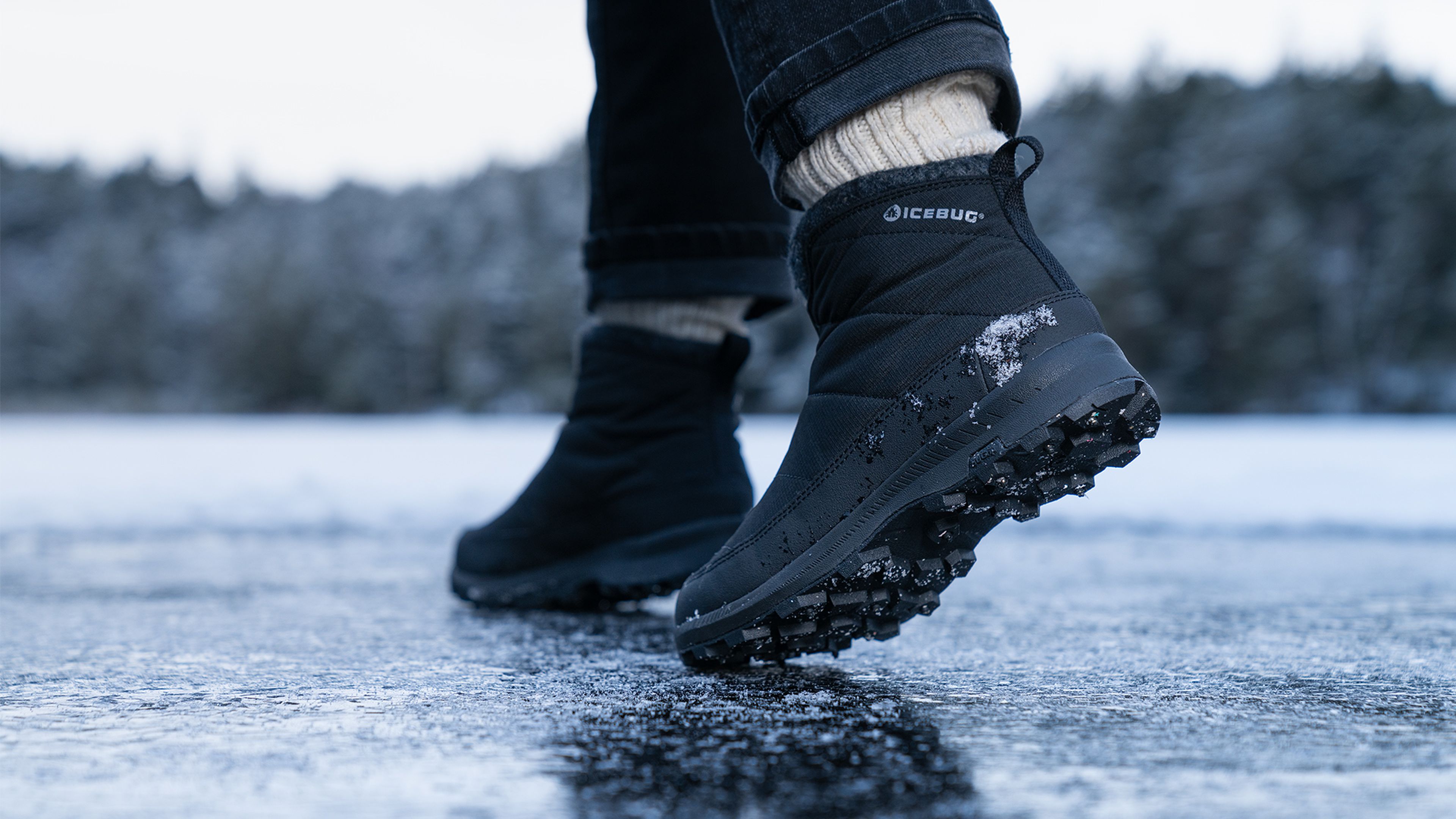 Person wearing black studded winter boots walking on ice, with snow on the soles, surrounded by a blurred snowy landscape.