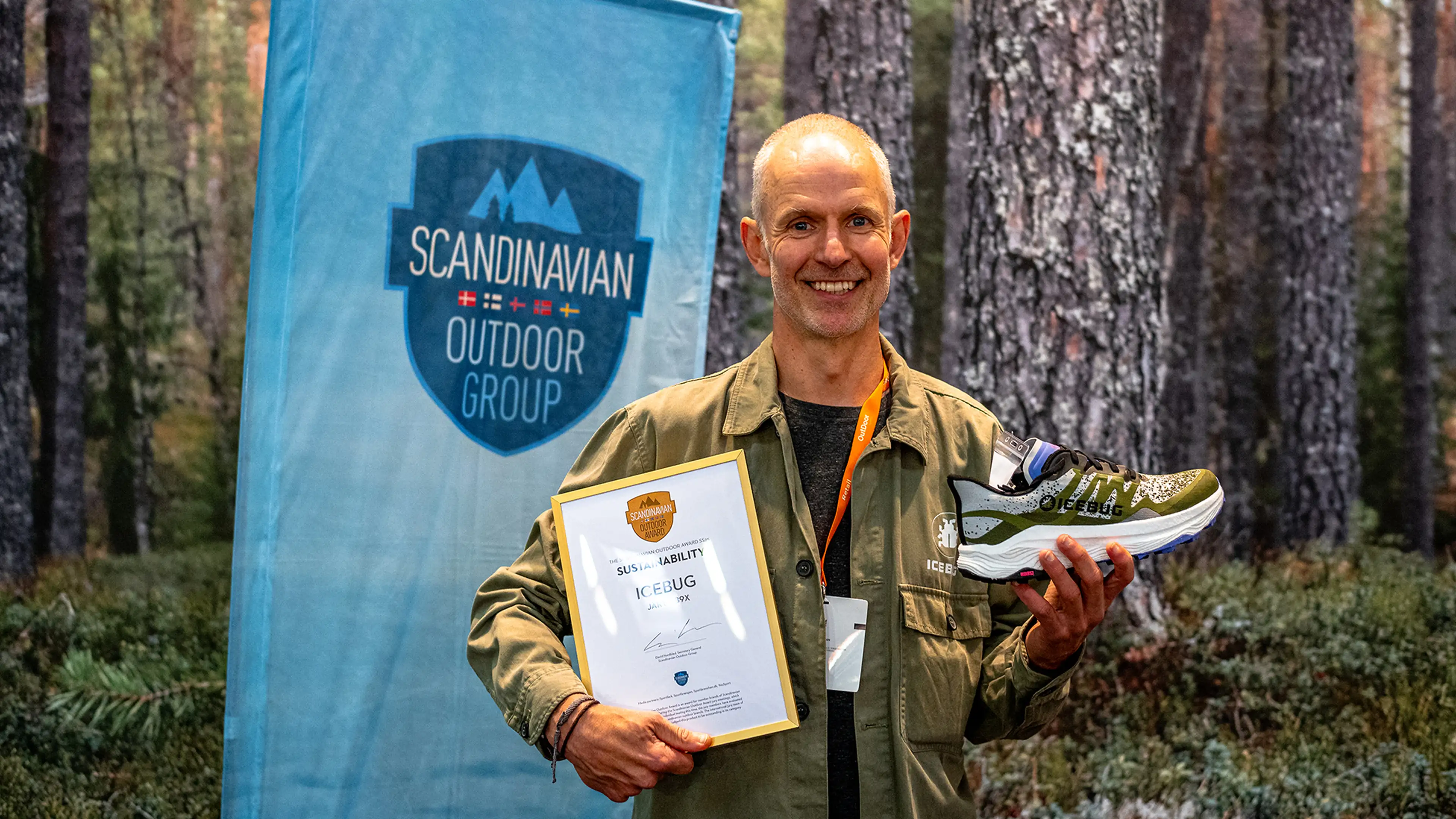 Man holding an award certificate and a shoe, standing in front of a banner that reads "Scandinavian Outdoor Group" in a forest setting.