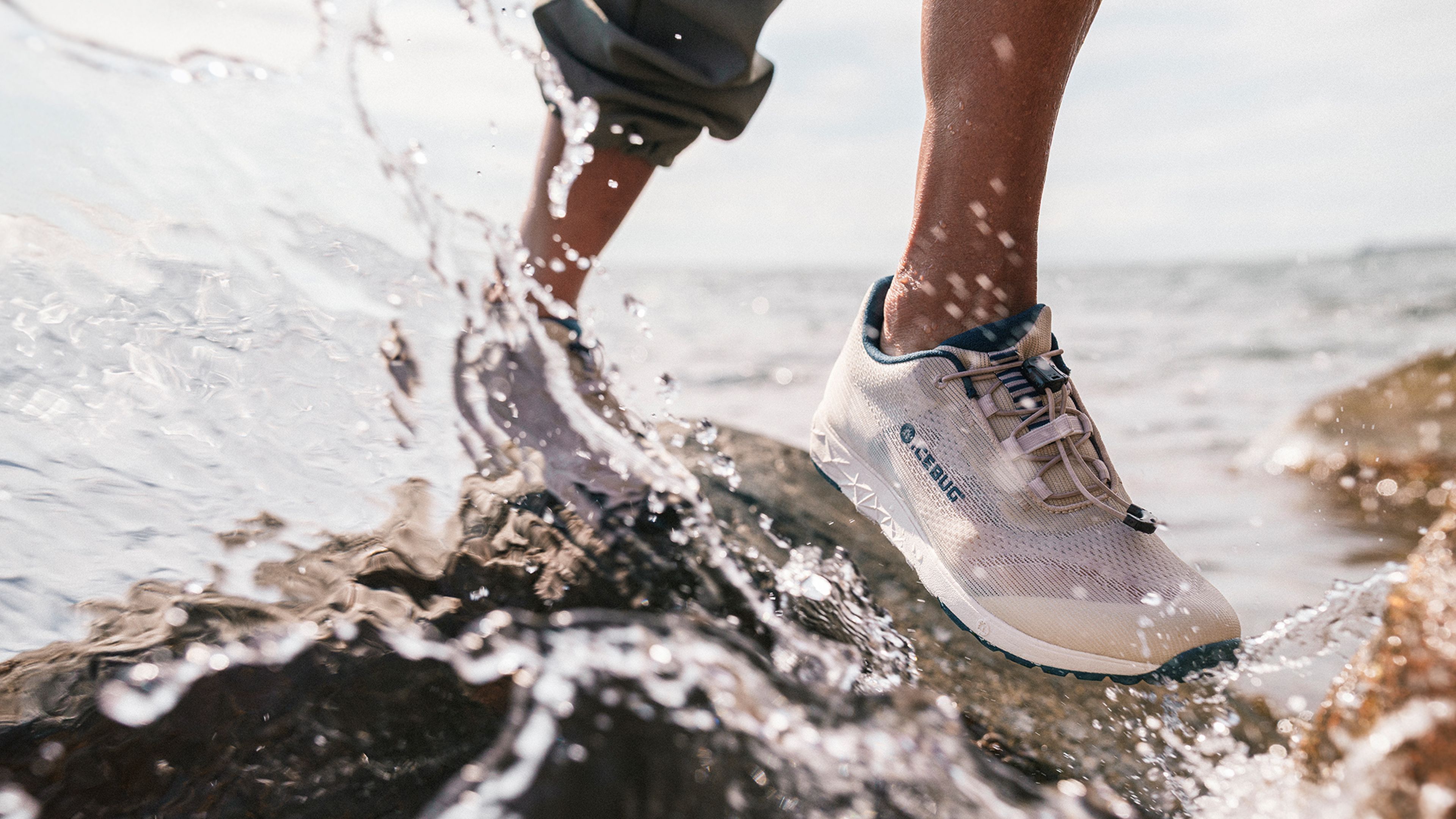 Person wearing white Icebug sneakers stepping on wet rocks near ocean waves.