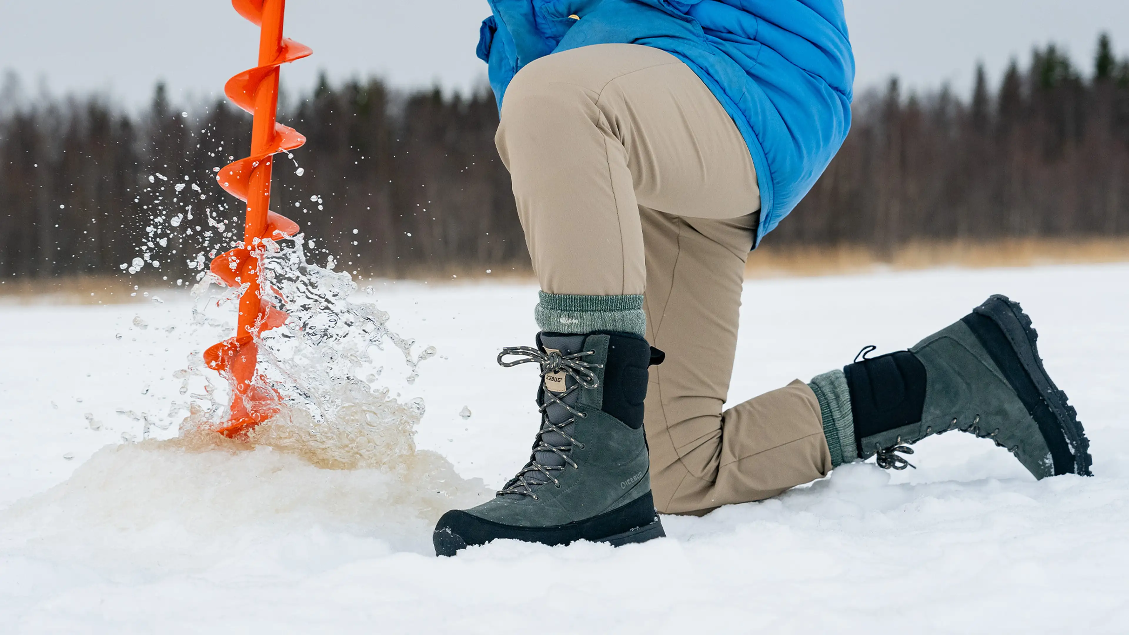 Person in winter clothing kneels on ice, using an orange auger to drill a hole, with water splashing around.