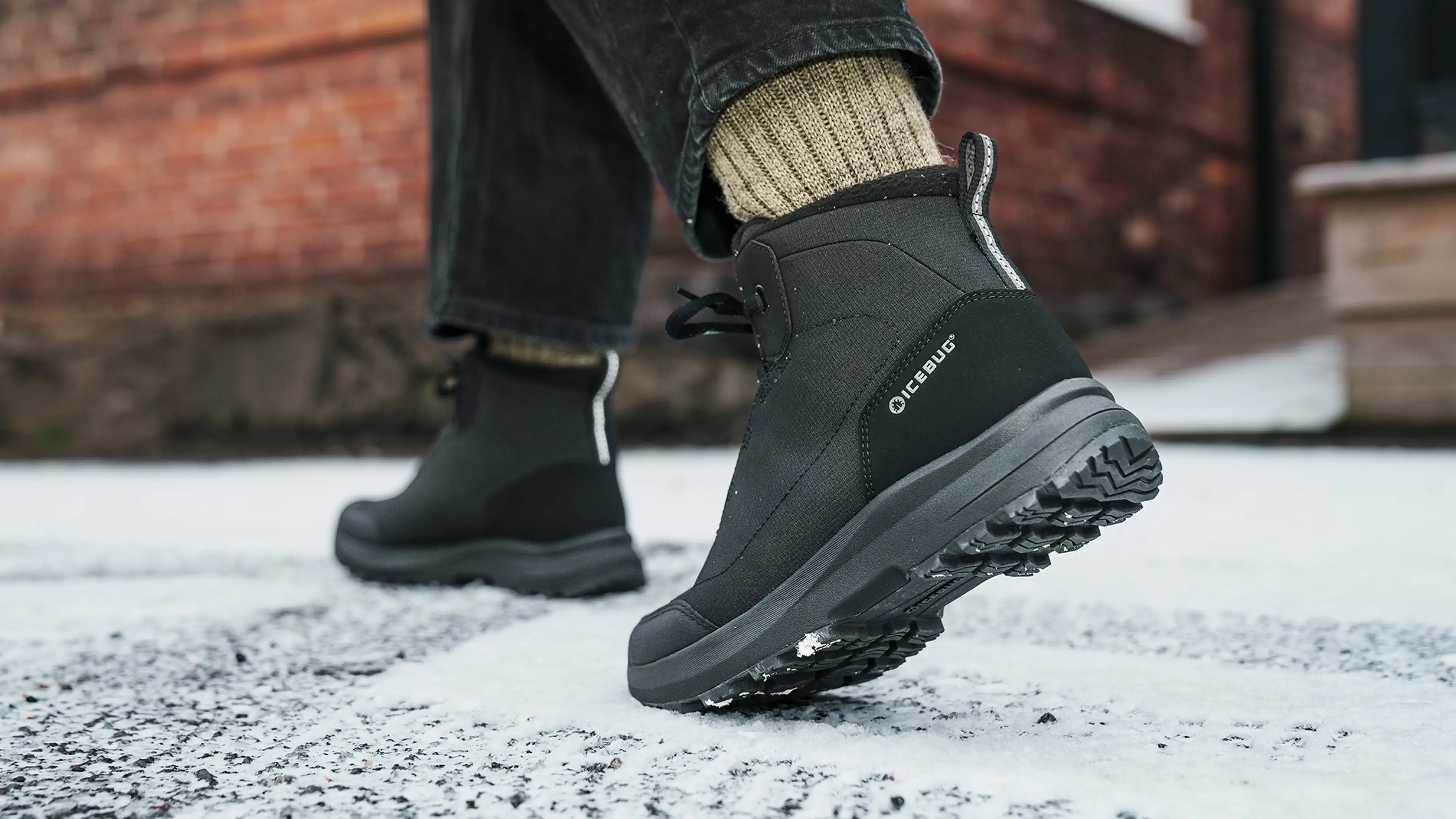 Close-up of a person wearing black winter boots walking on a snowy, icy pavement. The boots have a rugged sole for traction.