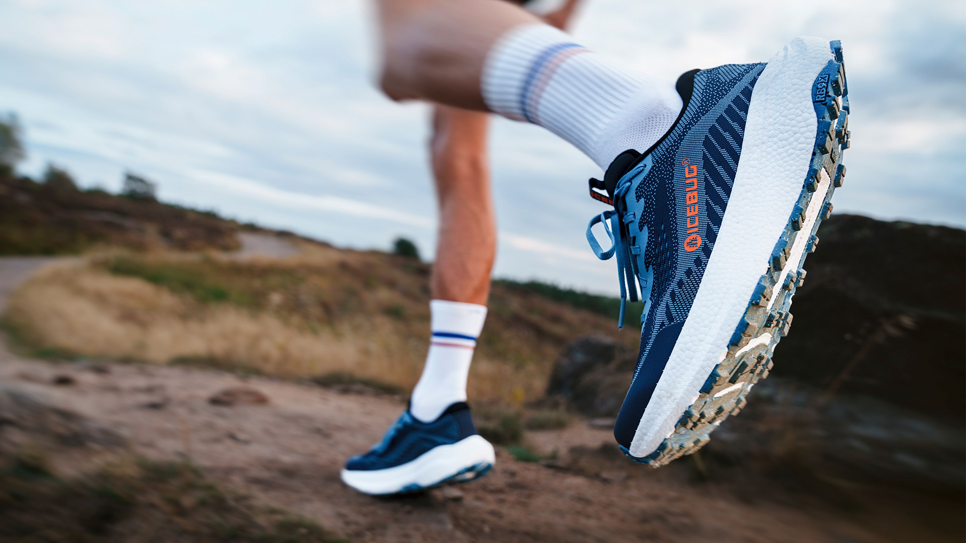 Close-up of a person running on a trail, wearing blue and white athletic shoes, with the background blurred, suggesting motion.