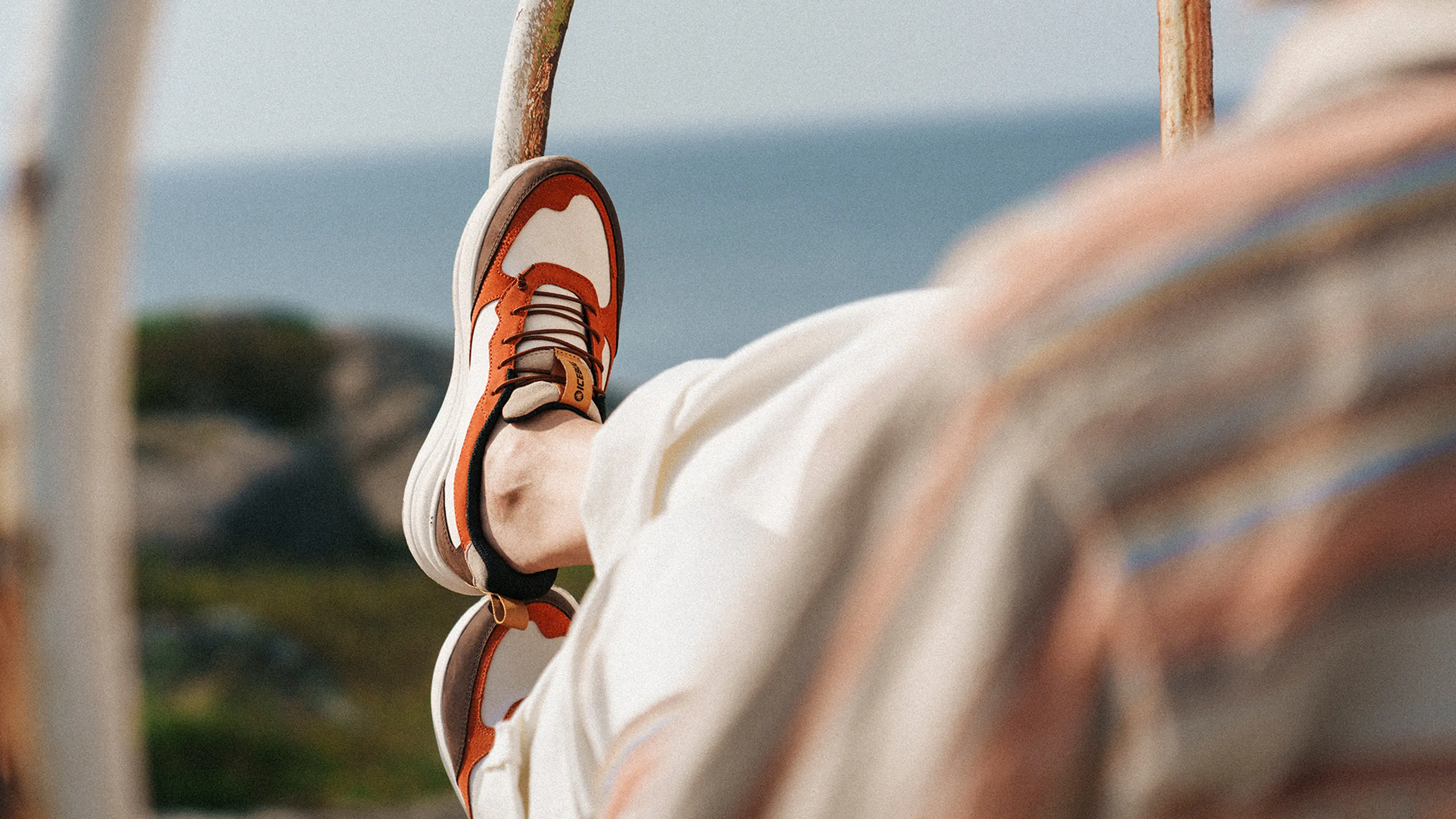 Person relaxing outdoors with feet up, wearing orange and white sneakers, overlooking a blurred natural landscape and sea.
