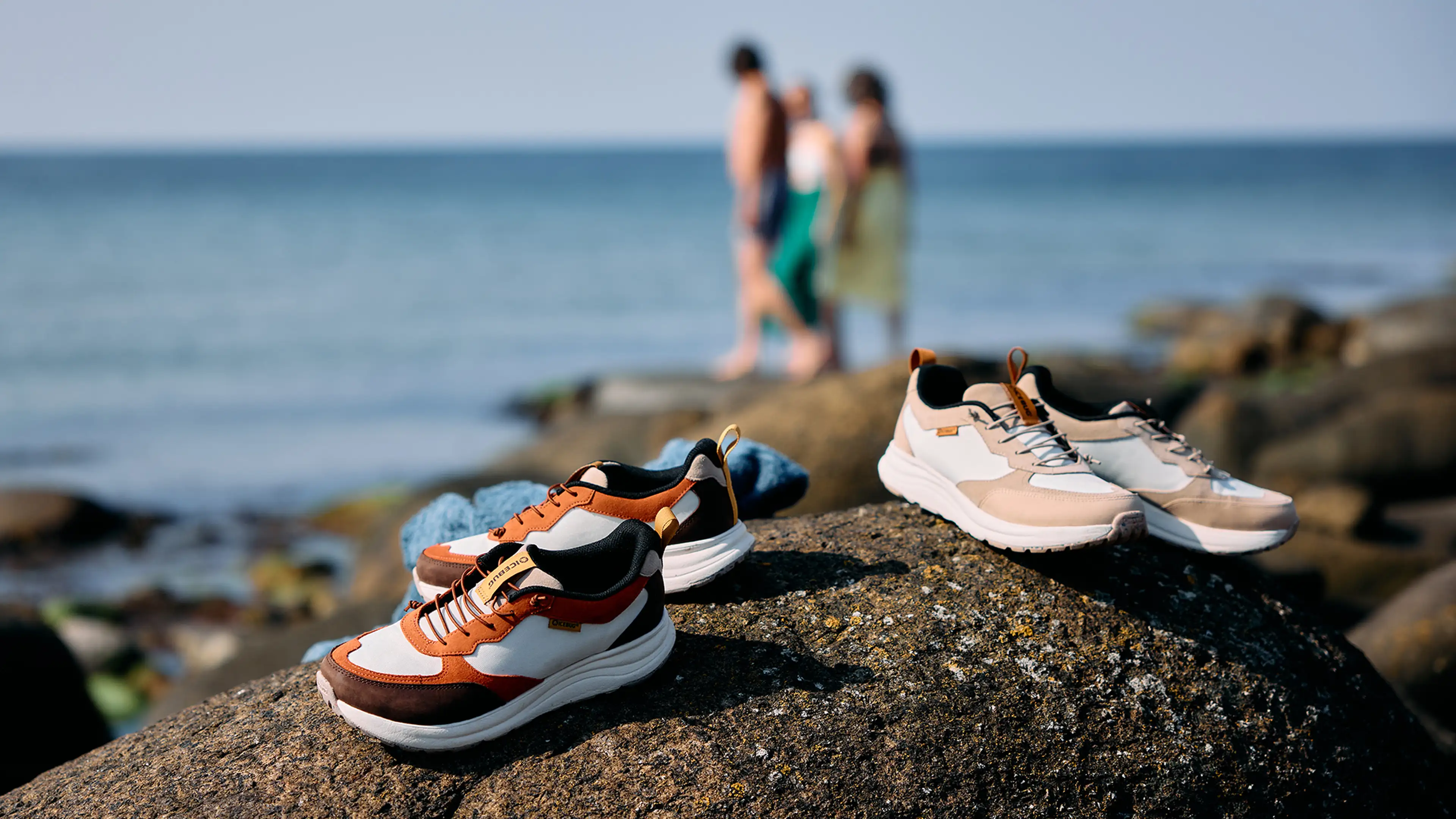 Two pairs of Icebug sneakers on a rock by the sea, with two blurred people in the background near the water's edge.