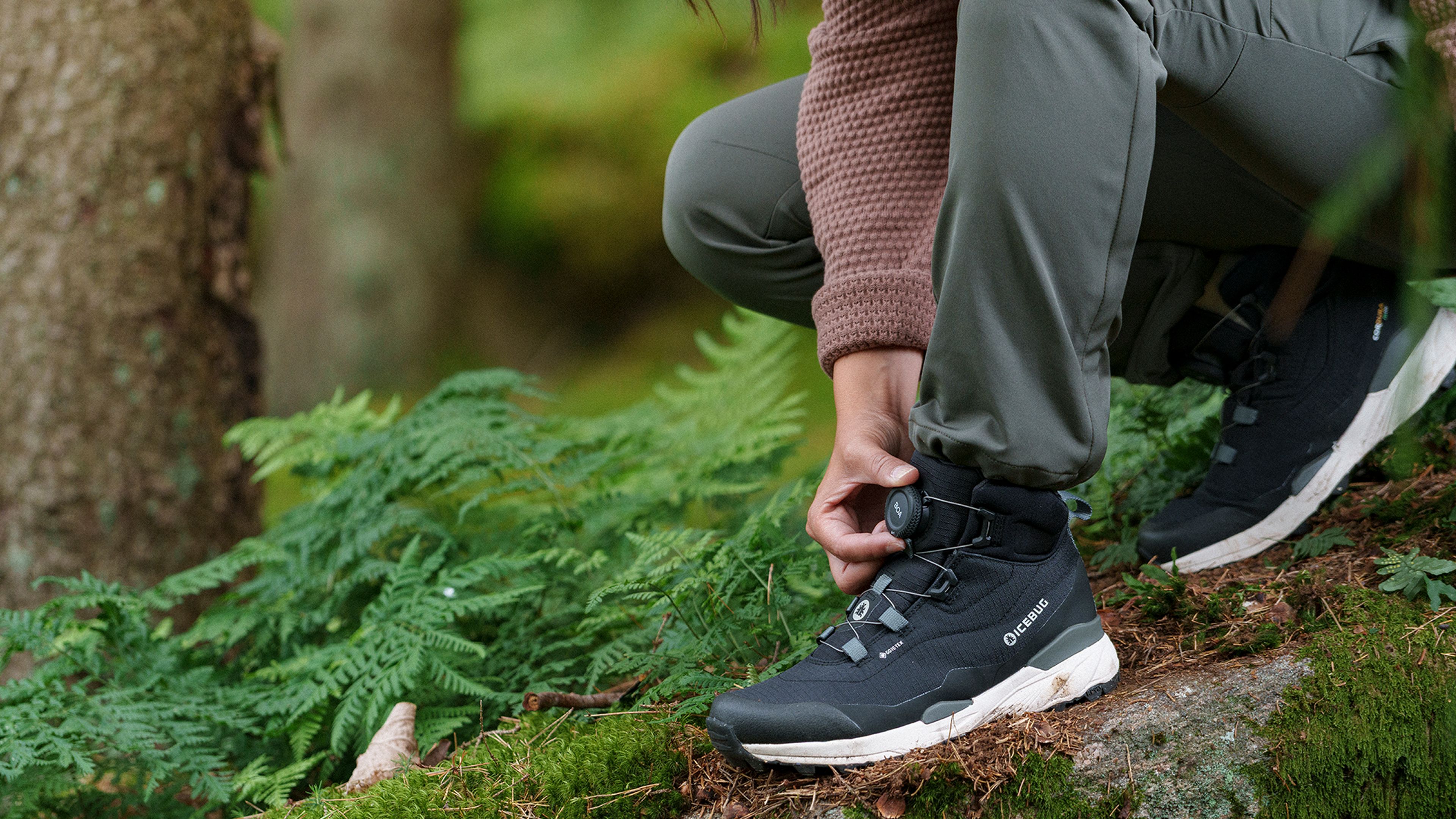 Person tying a black hiking boot in a lush, green forest setting, wearing gray pants and a brown sweater.