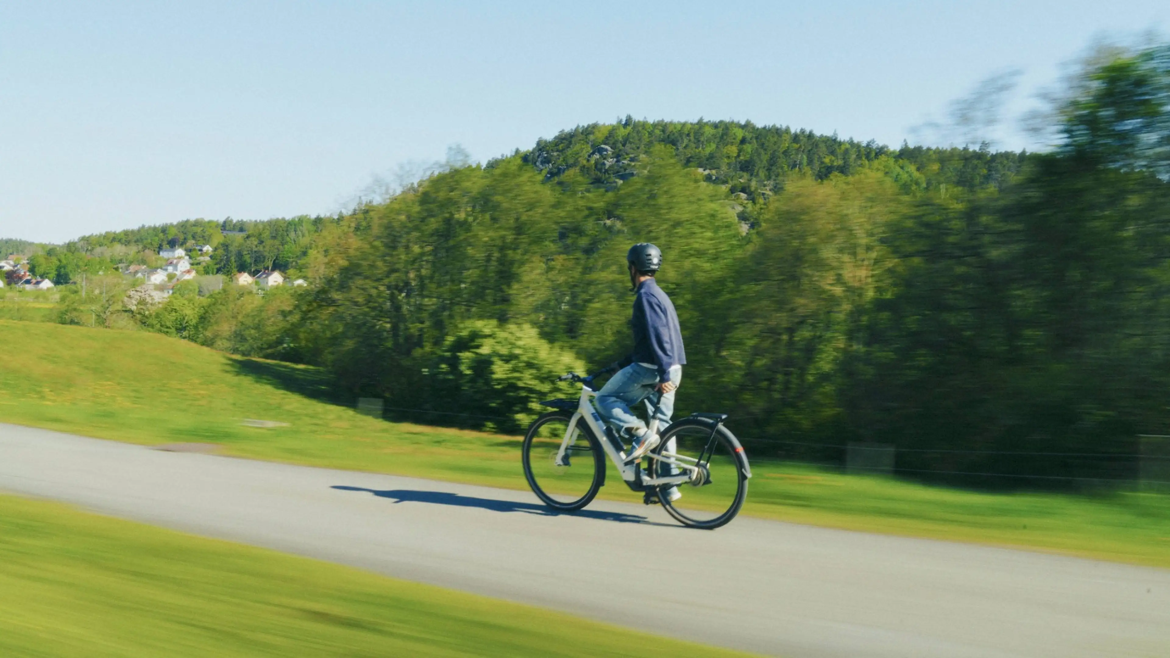 Person cycling on a road through a scenic, lush green landscape with rolling hills under a clear blue sky.