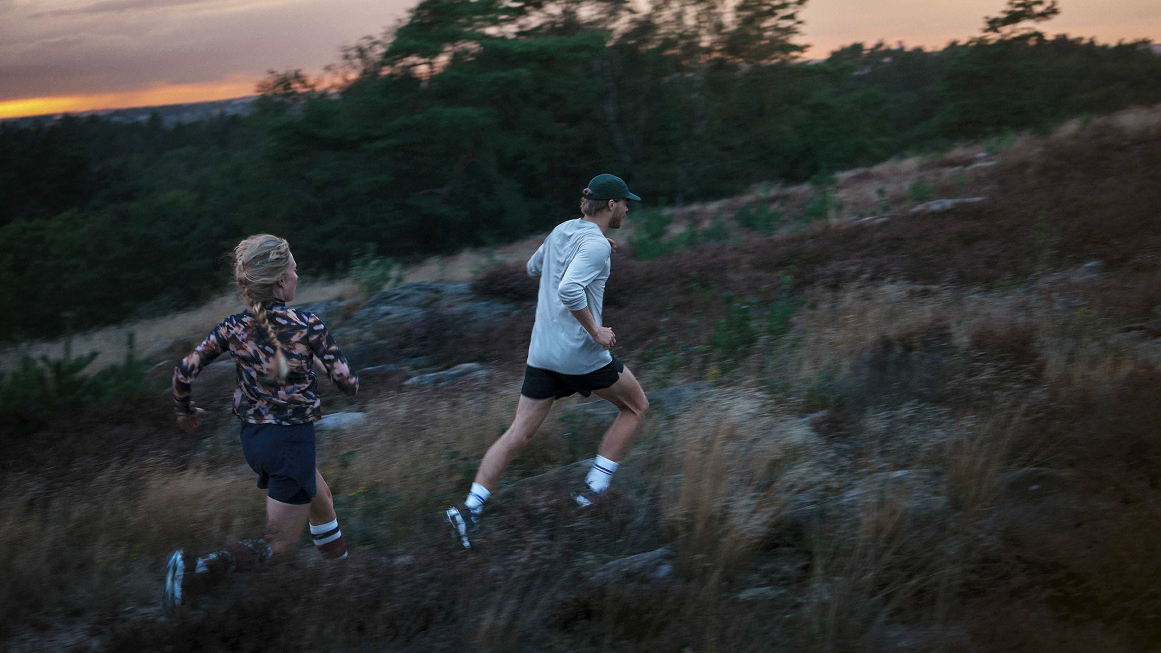 Two people in athletic gear trail running through a rugged, grassy landscape at sunset, with trees and a colorful sky in the background.