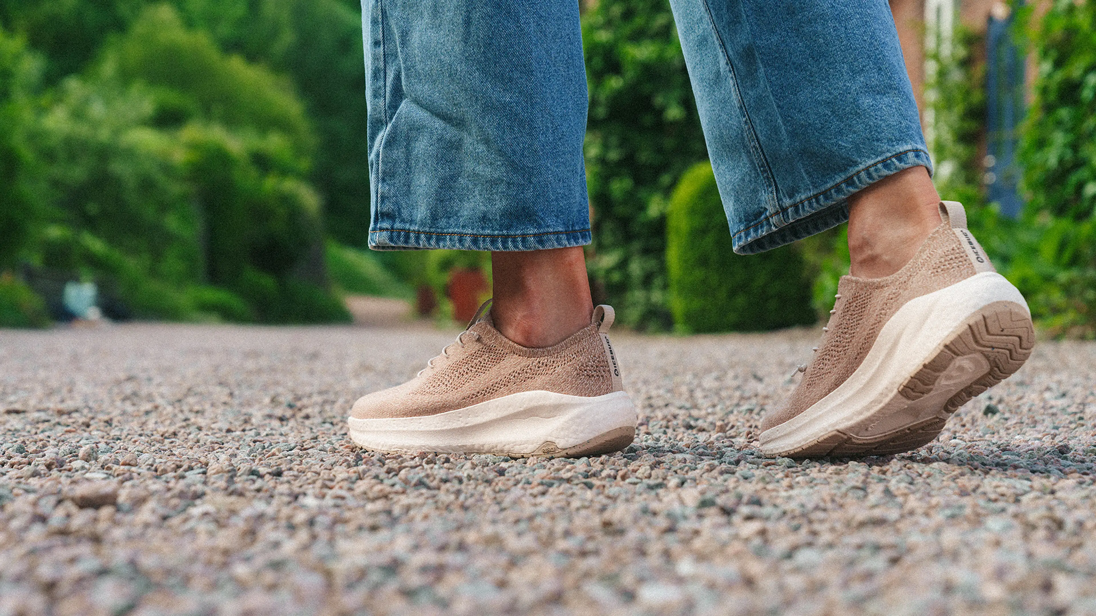 Close-up of a person wearing beige sneakers and blue jeans, walking on a gravel path surrounded by greenery.