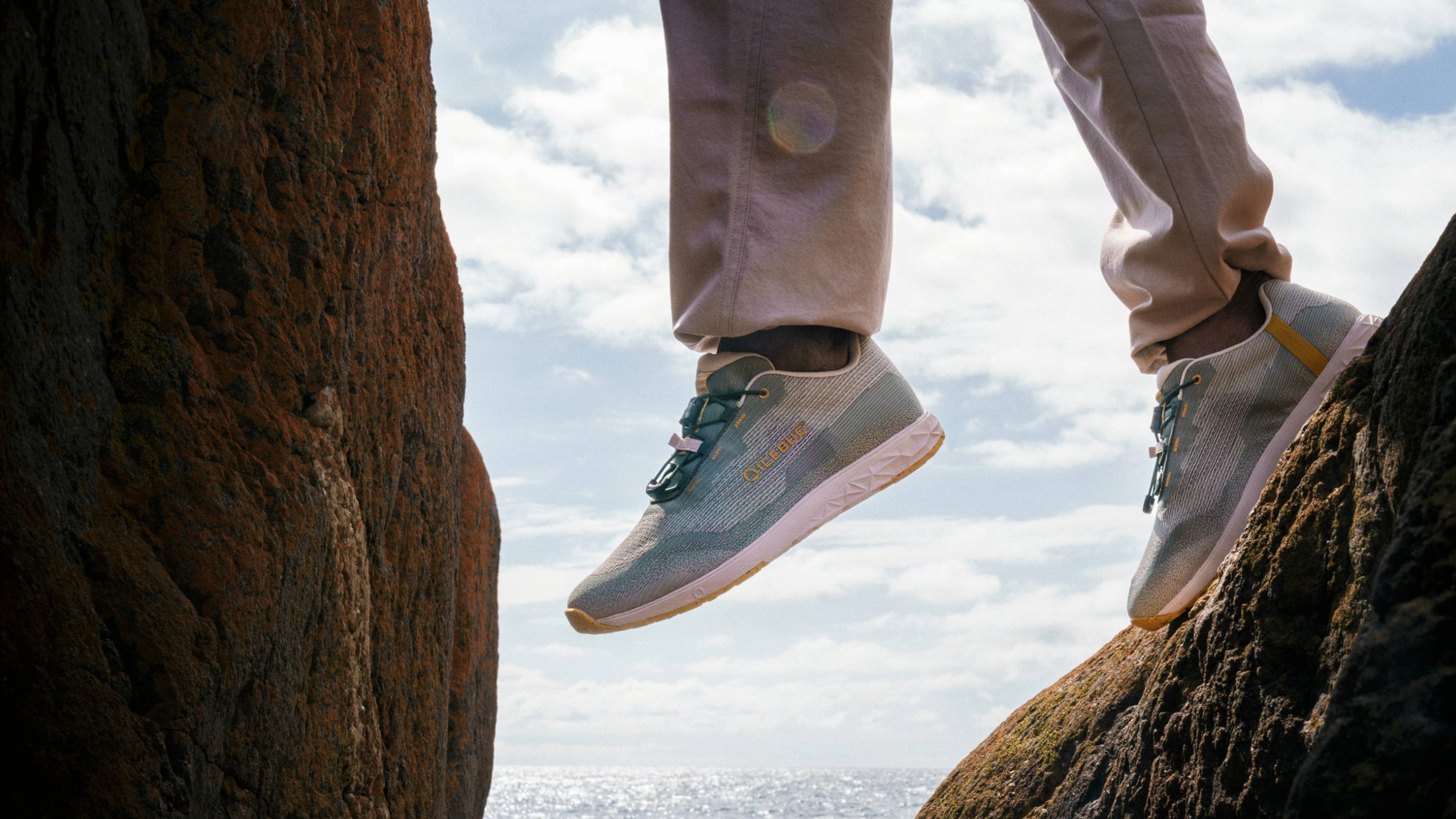 Person wearing light gray sneakers and beige pants standing between rocky cliffs with the ocean and a cloudy sky in the background.