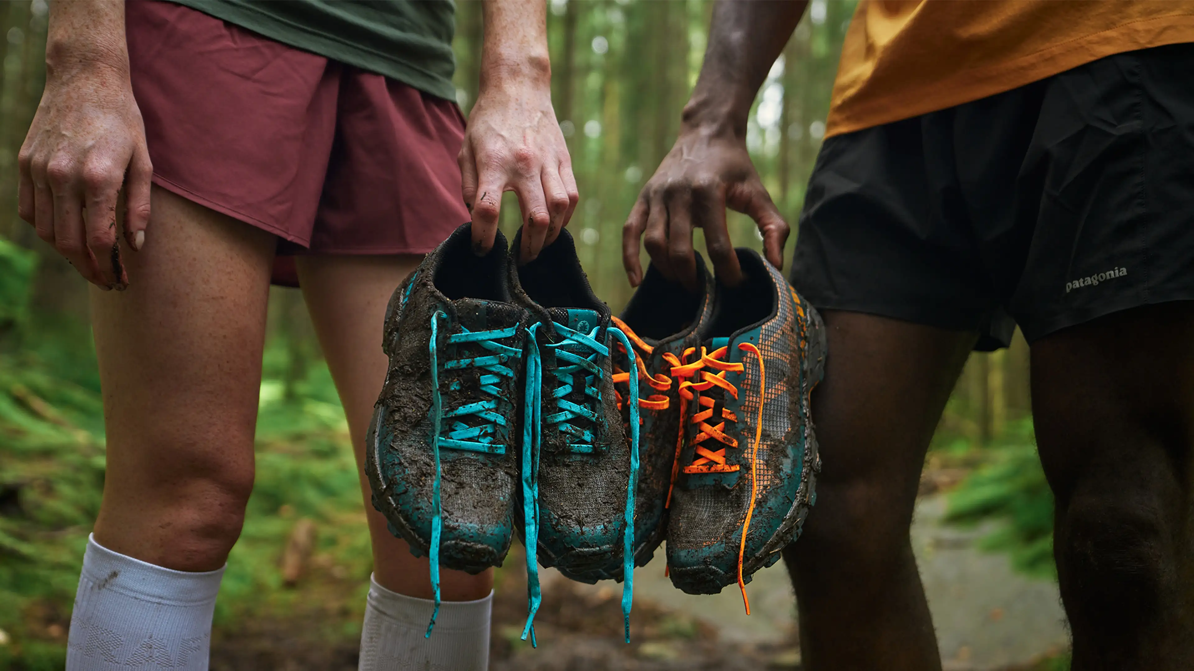 Two people holding muddy trail running shoes with colorful laces in a forest setting, wearing shorts and standing on a dirt path.