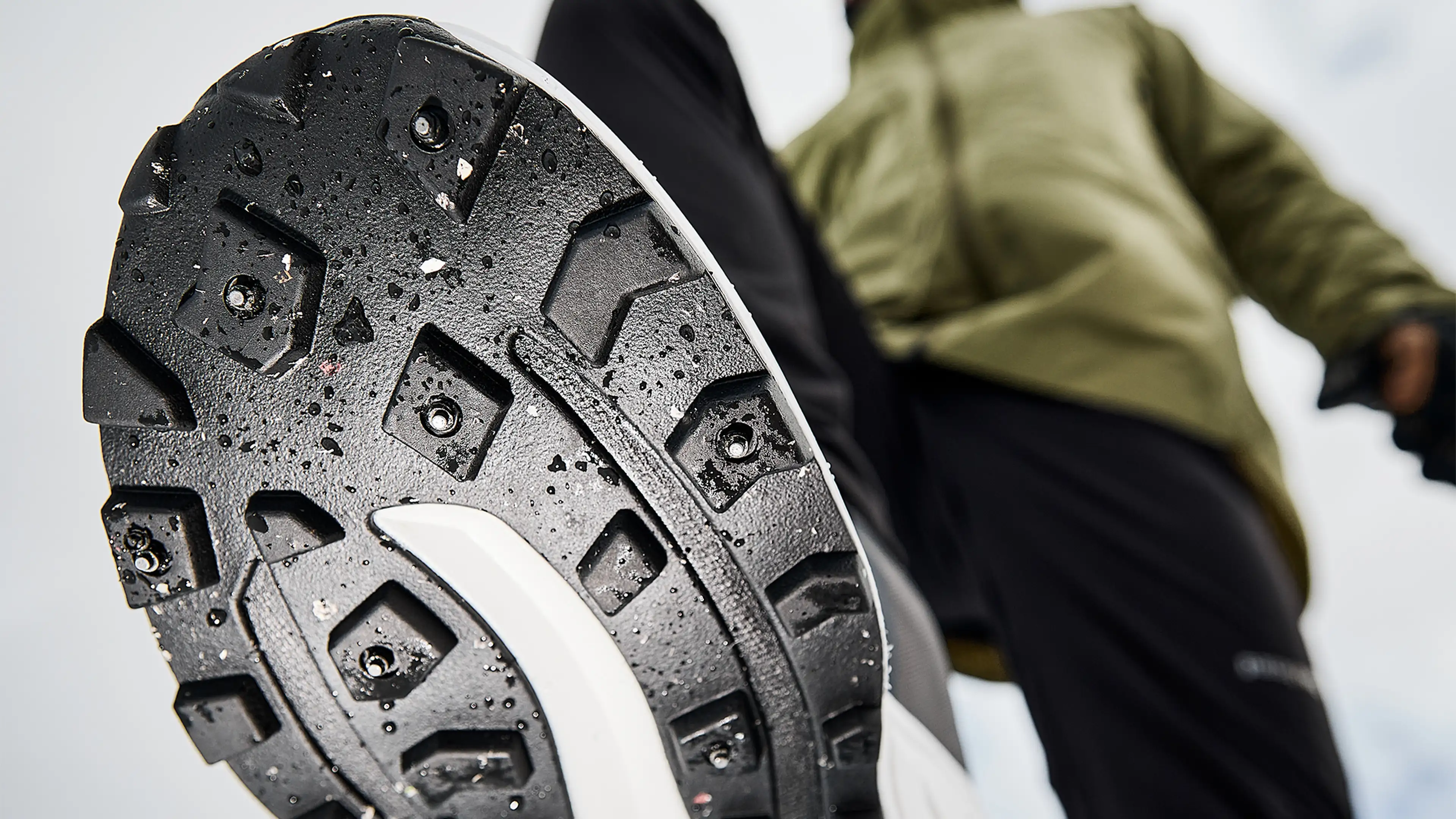 Close-up of a rugged, dirt-speckled shoe sole with spikes, worn by a person in outdoor clothing, suggesting a running or outdoor activity.