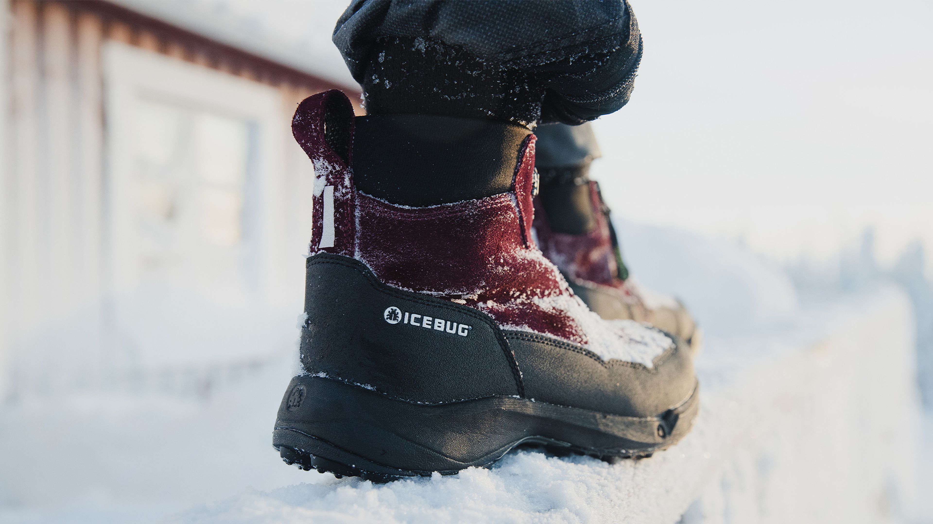 Close-up of a person wearing red and black Icebug boots, standing on a snowy surface near a building.