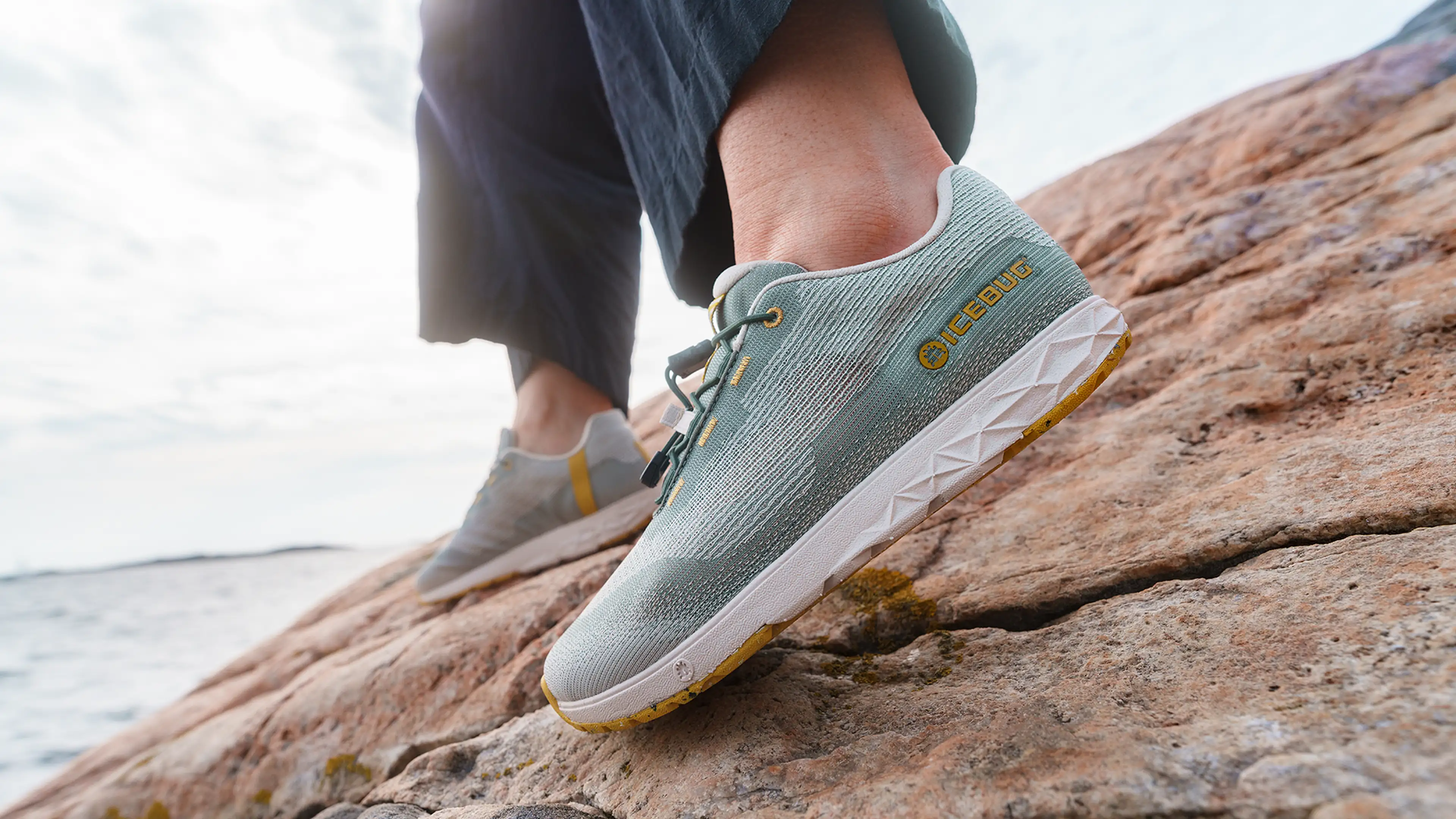 Close-up of a person wearing light green athletic Icebug shoes with white soles, standing on a rocky surface by the water under a cloudy sky.