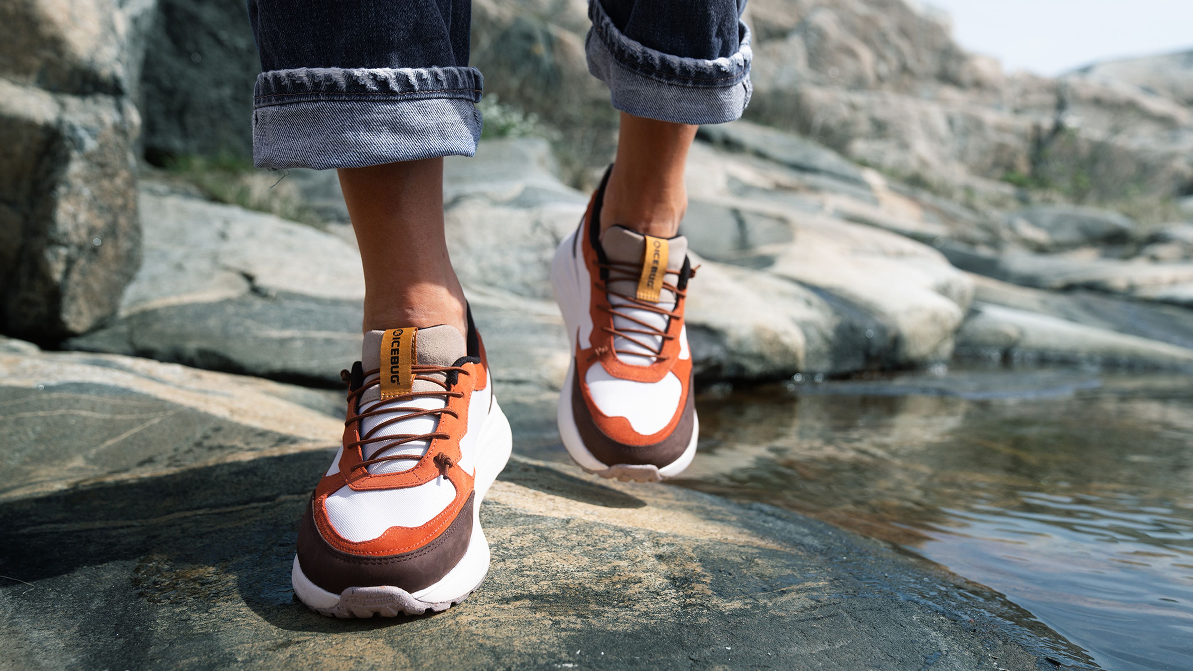 Person wearing orange and white sneakers walking on rocks by water, with jeans rolled up.