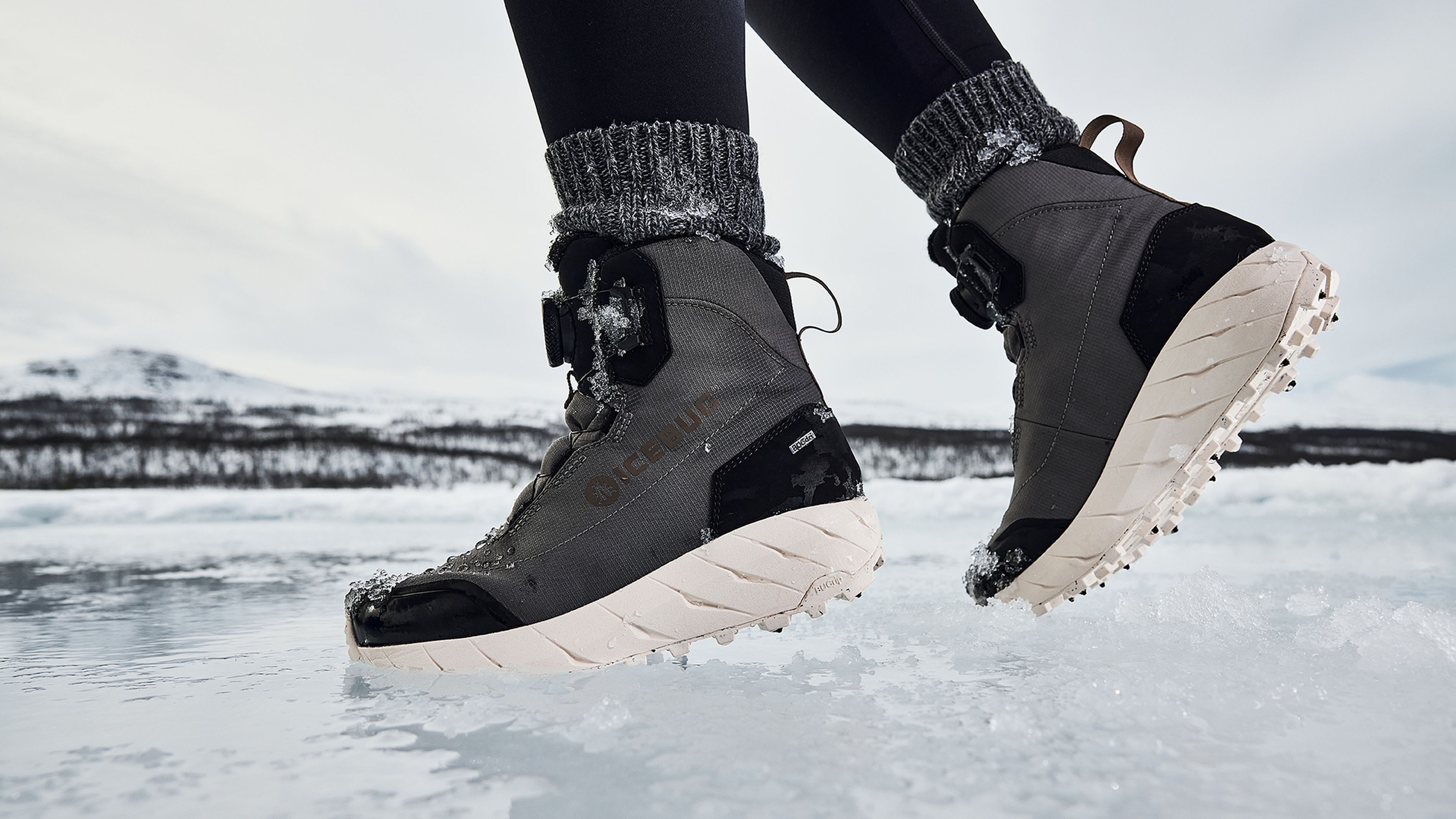 Close-up of a person wearing gray winter boots with thick soles, walking on ice. Snowy mountains and cloudy sky in the background.