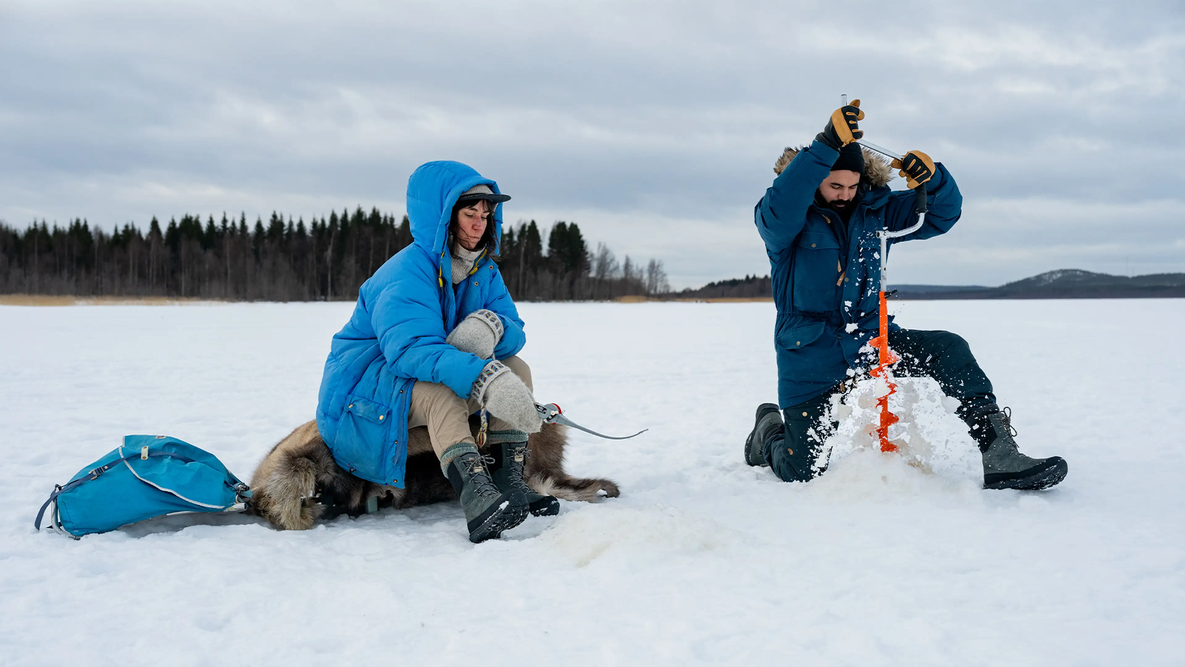 Two people in winter clothing are ice fishing on a frozen lake. One is drilling a hole while the other sits nearby with a dog. Forest in the background.