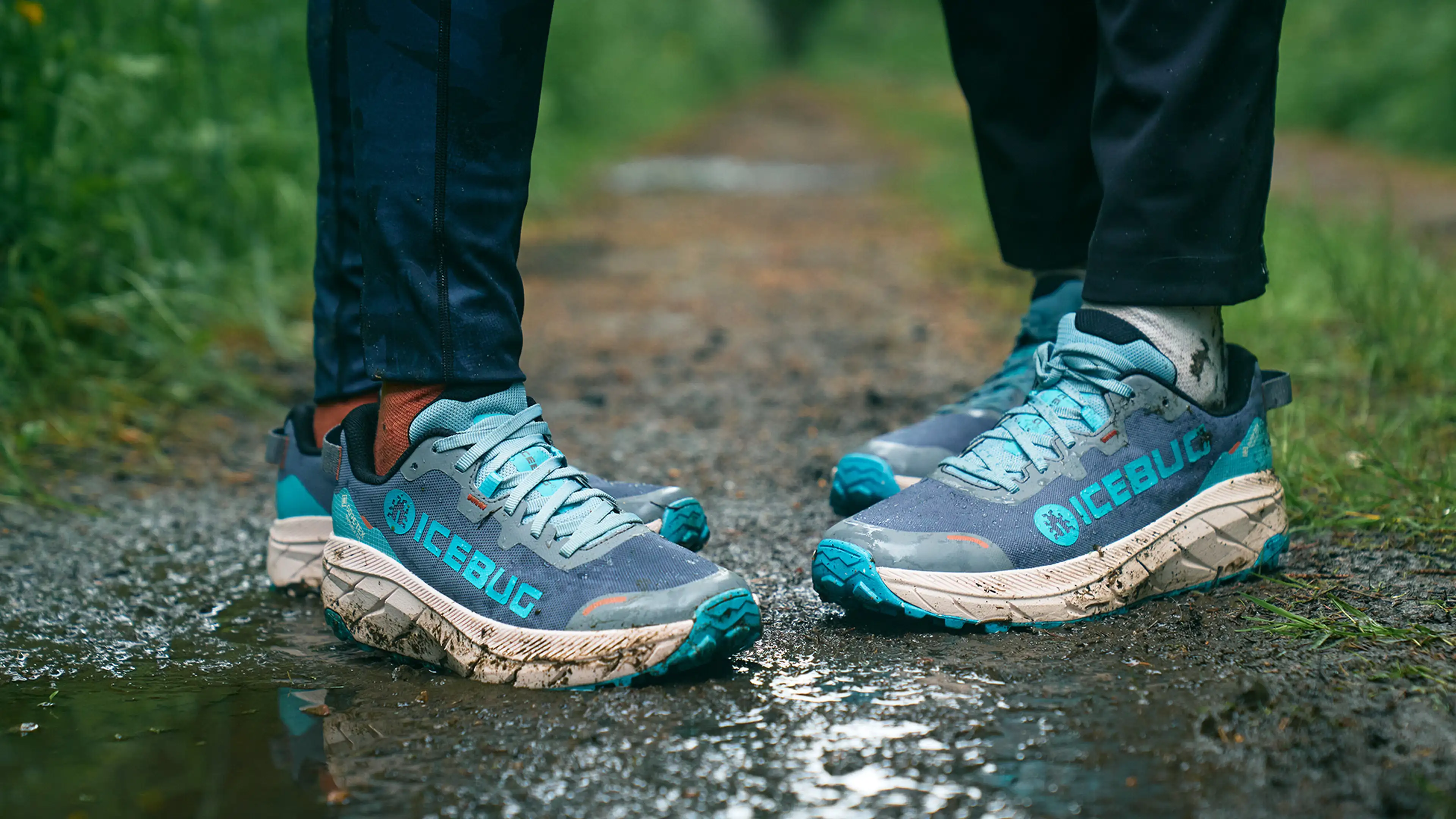 Two people wearing blue and turquoise trail running shoes stand on a muddy path, with grass and trees in the background.