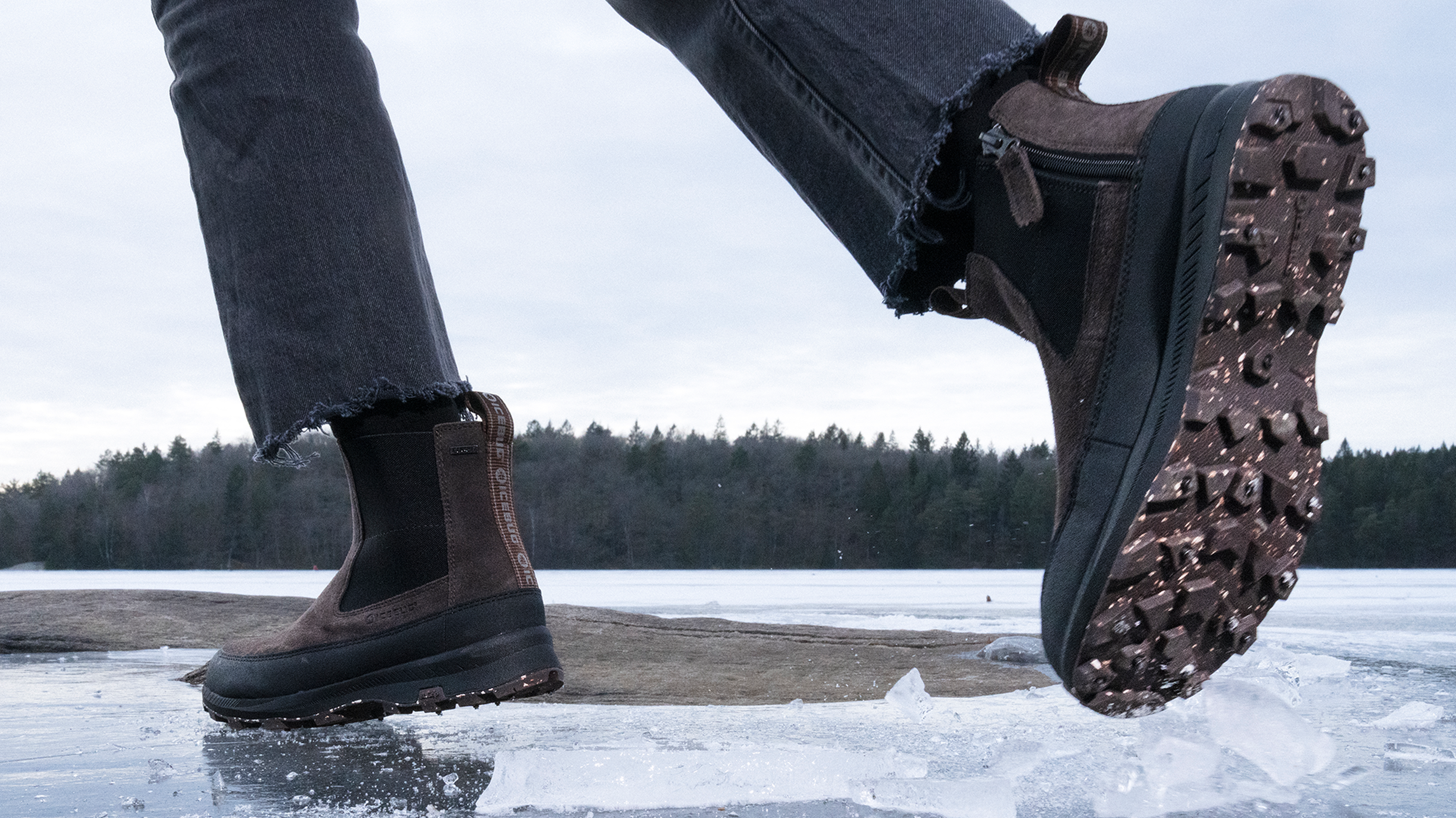 Close-up of a person walking on ice, wearing brown studded winter boots with thick soles and spikes. The background shows a frozen lake and trees.