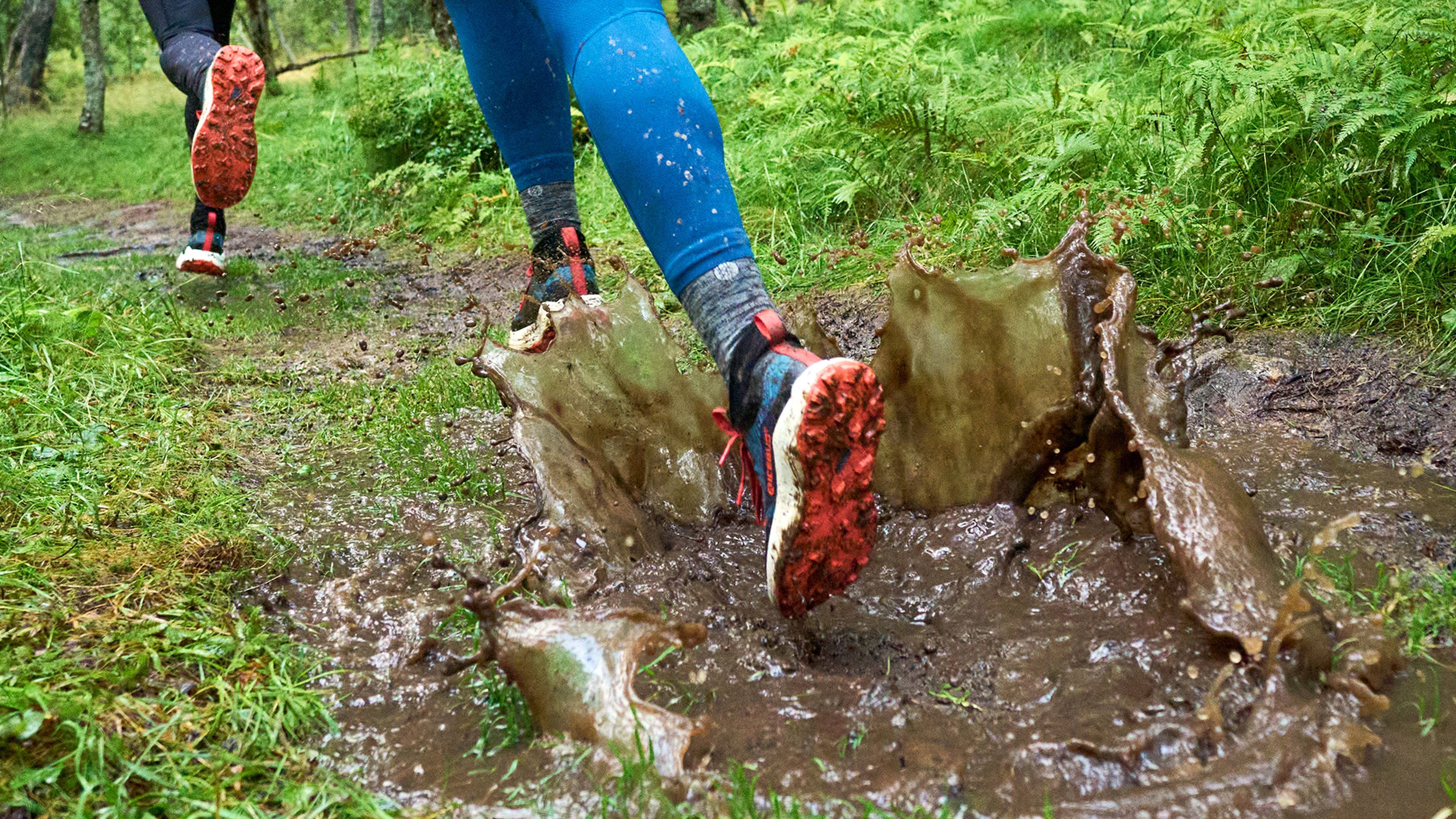 Runners splash through muddy puddles on a forest trail, wearing bright blue leggings and oranged-soled shoes.