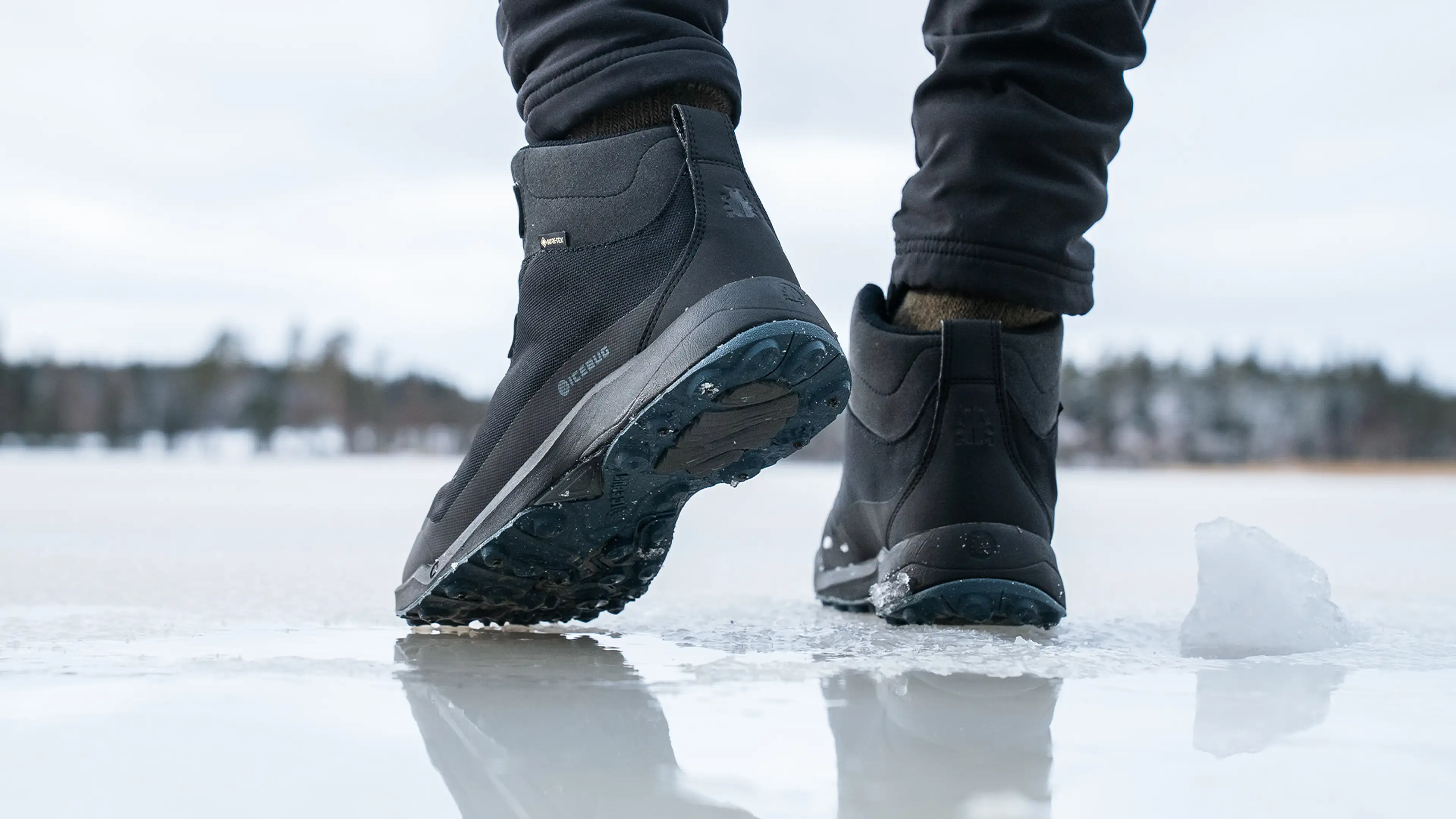 Close-up of a person wearing black waterproof boots walking on an icy surface, with a snowy landscape in the background.