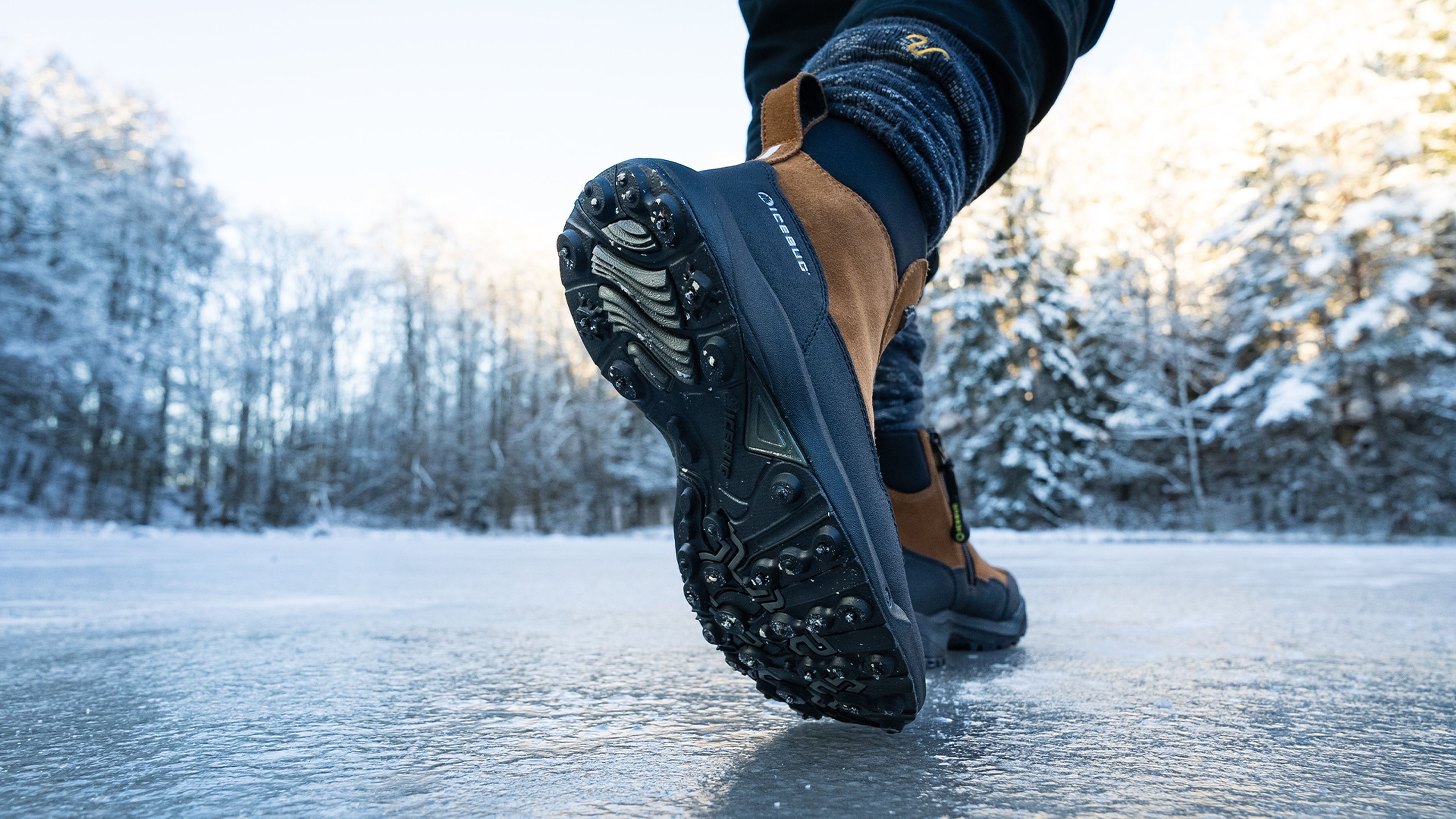 Close-up of a person walking on ice, wearing brown and black winter boots with studded soles. Snow-covered trees are in the background.