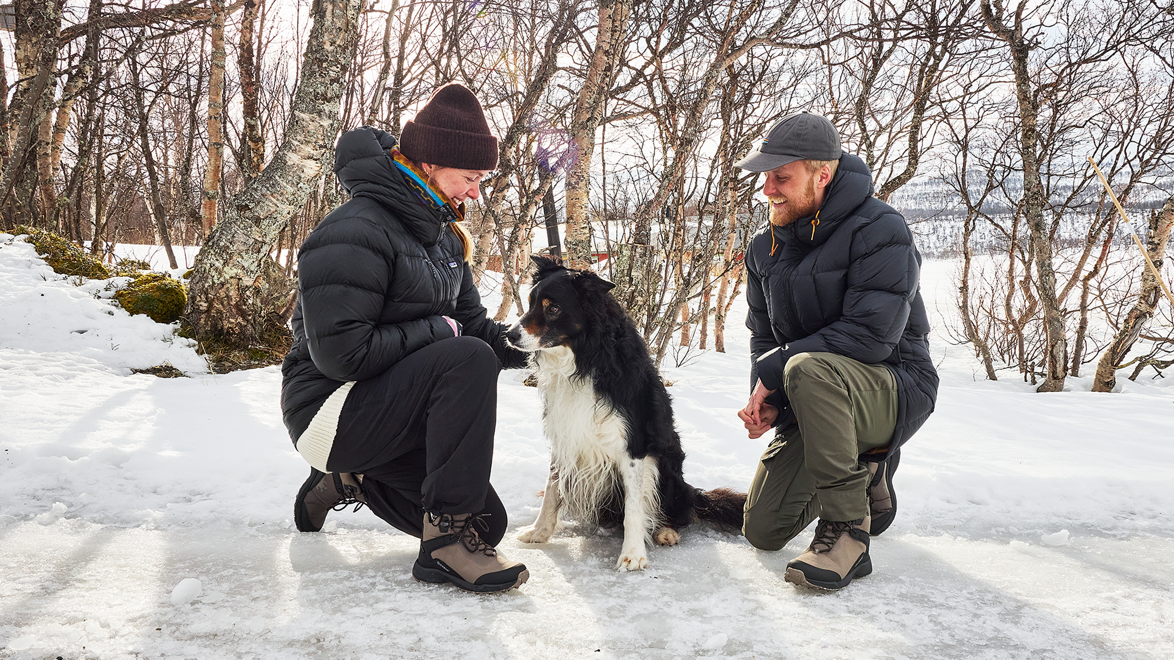 A man and woman in winter clothing kneel beside a black and white dog on a snowy path, surrounded by bare trees.