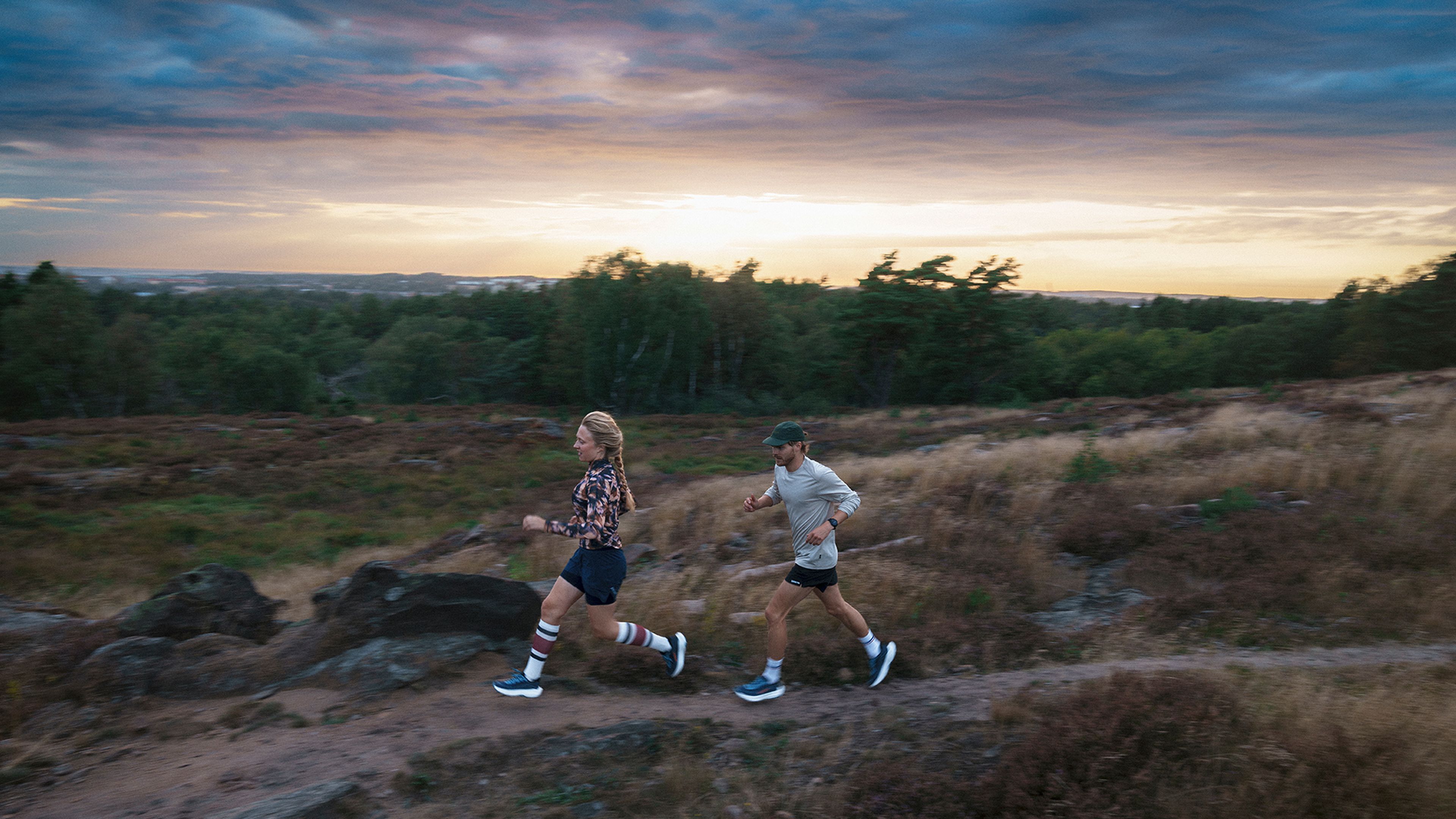 Two people running on a trail through a grassy landscape during a vibrant sunset, with trees and a dramatic sky in the background.