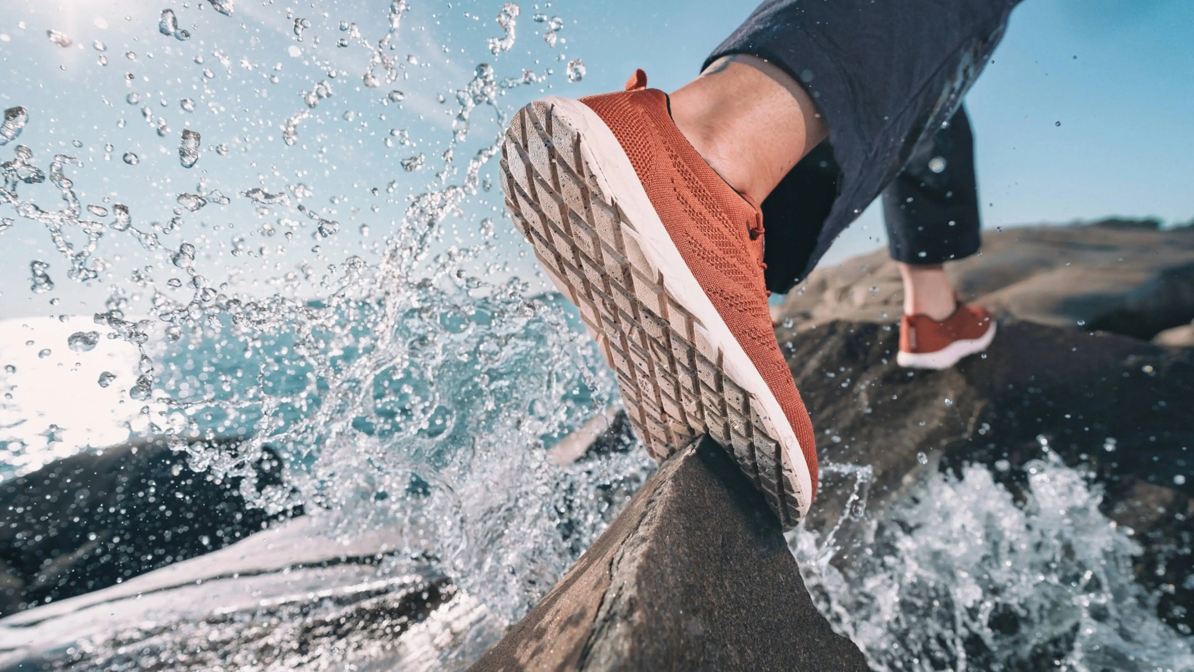 Close-up of a person wearing red sneakers stepping on a wet rock, with water splashing around, under a bright blue sky.