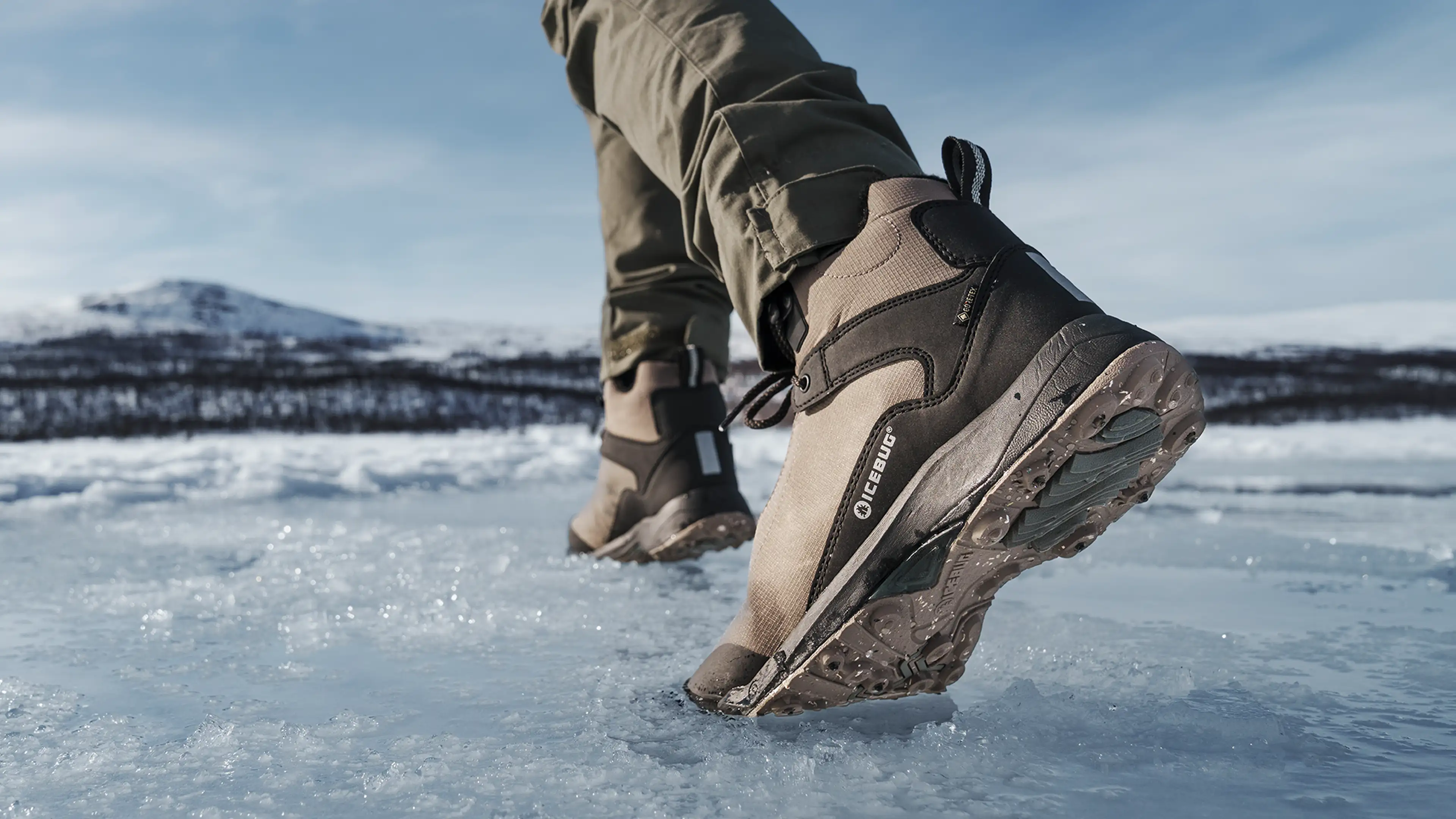 Close-up of a person wearing brown hiking boots walking on icy terrain, with snow-covered mountains in the background under a clear blue sky.