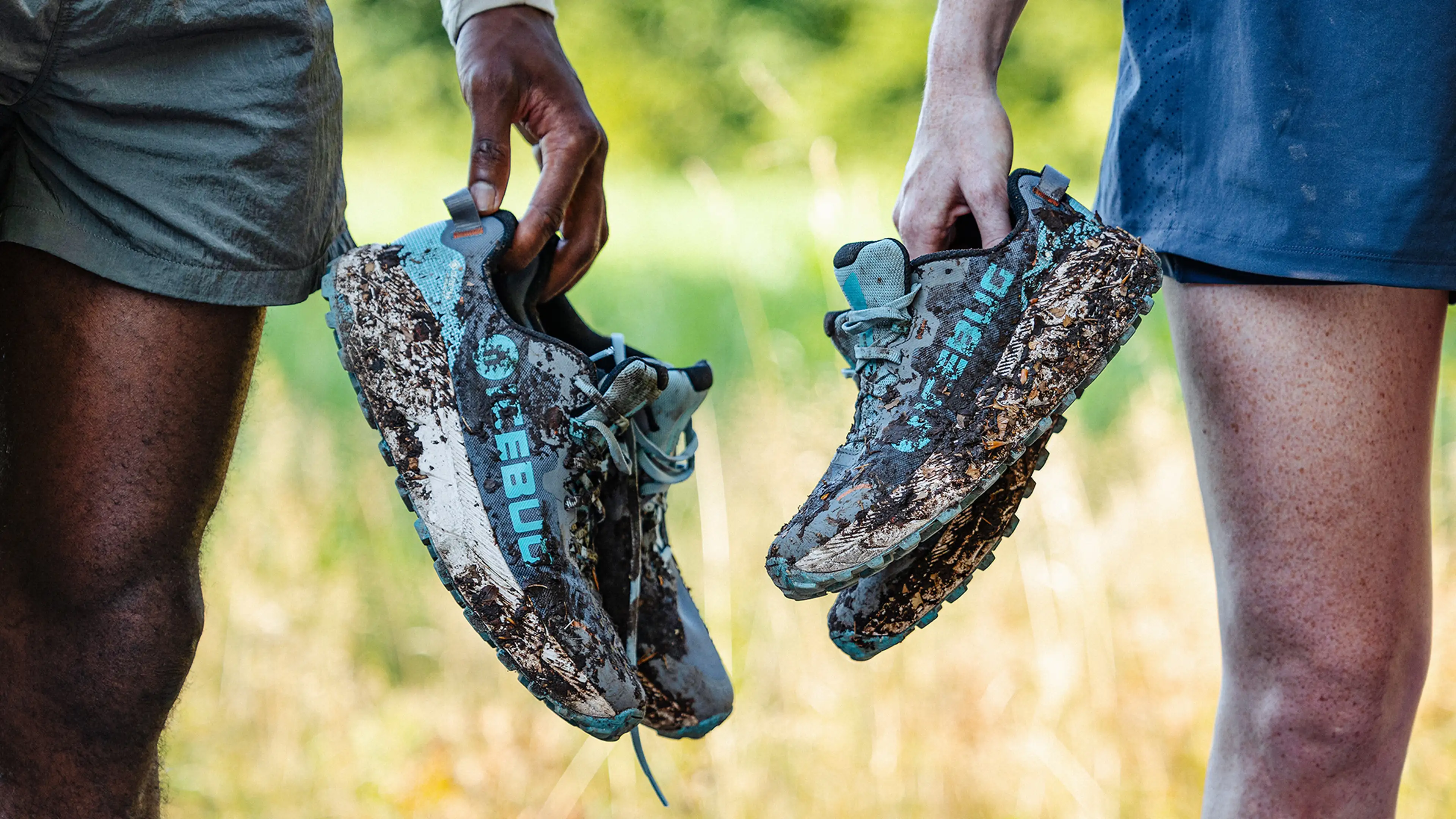Two people holding mud-covered athletic shoes outdoors, with grass and trees in the background. One person wears shorts; the other has brown skin.