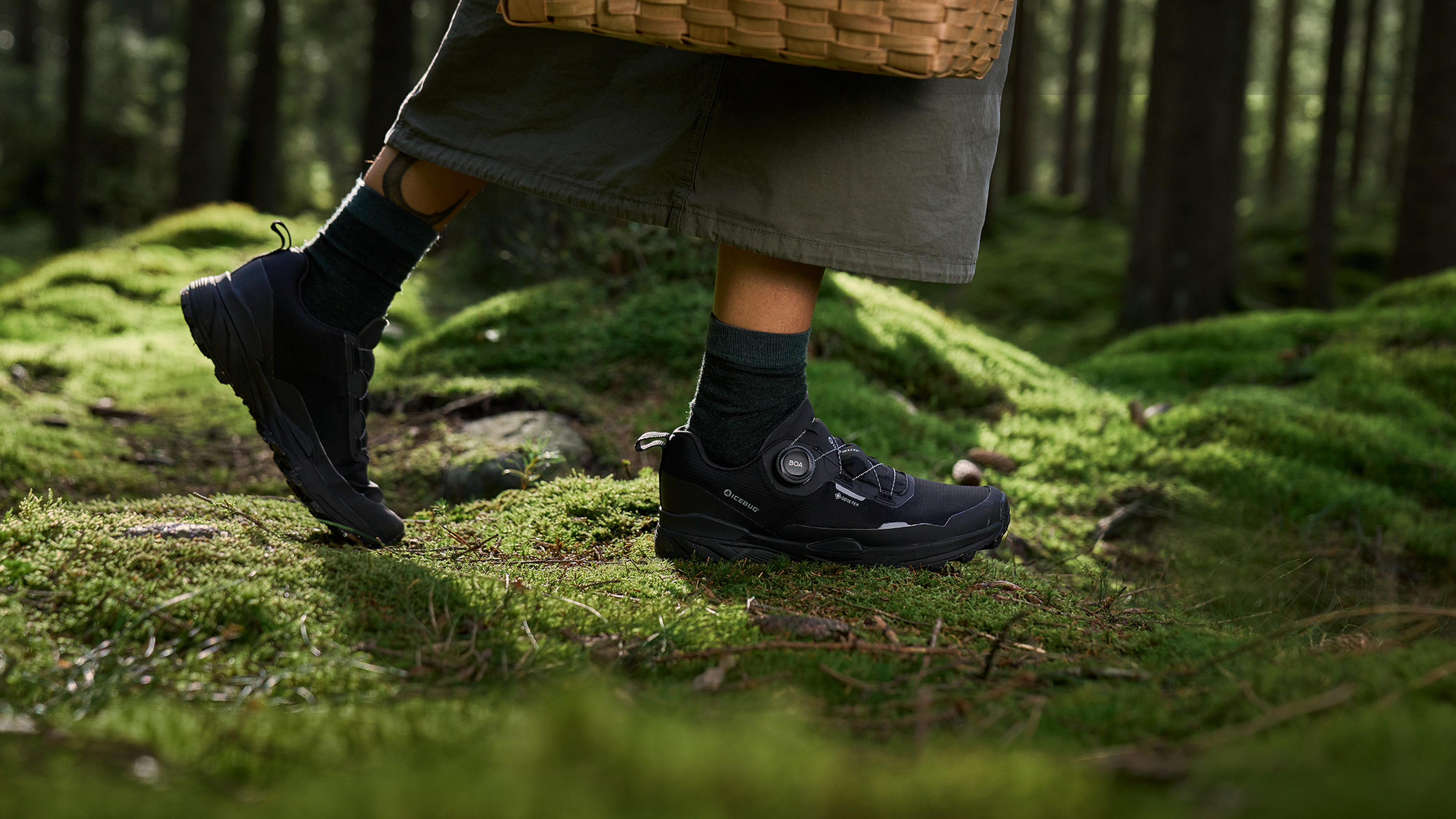 A person in black hiking shoes and green socks walks on a mossy forest floor, carrying a woven basket.