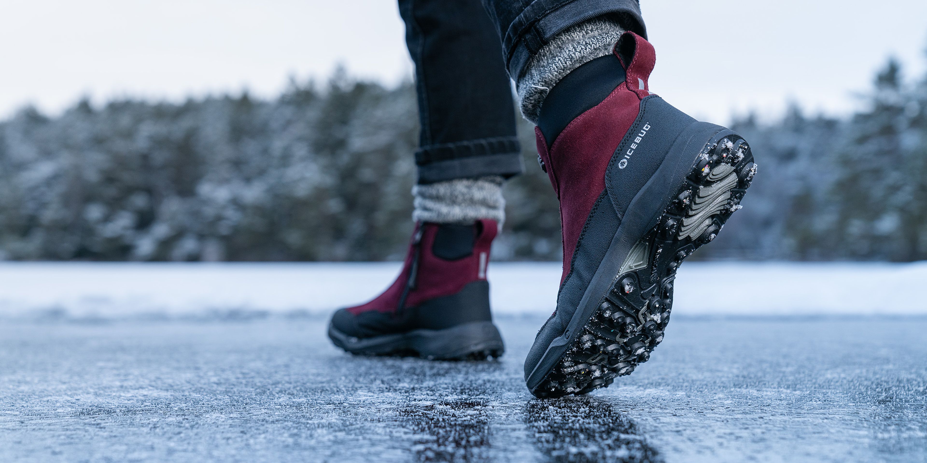 Person walking on icy ground wearing red winter boots with sturdy soles, surrounded by a snowy landscape and trees in the background.