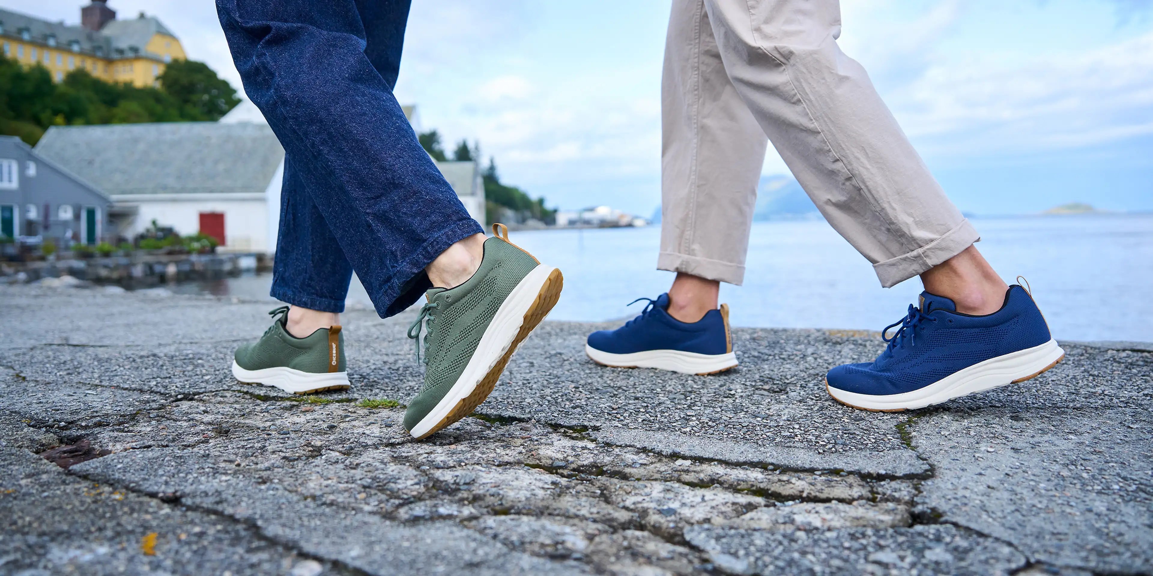 Two people walking on a seaside path, wearing casual sneakers—one green, one blue. Background features houses and a cloudy sky.