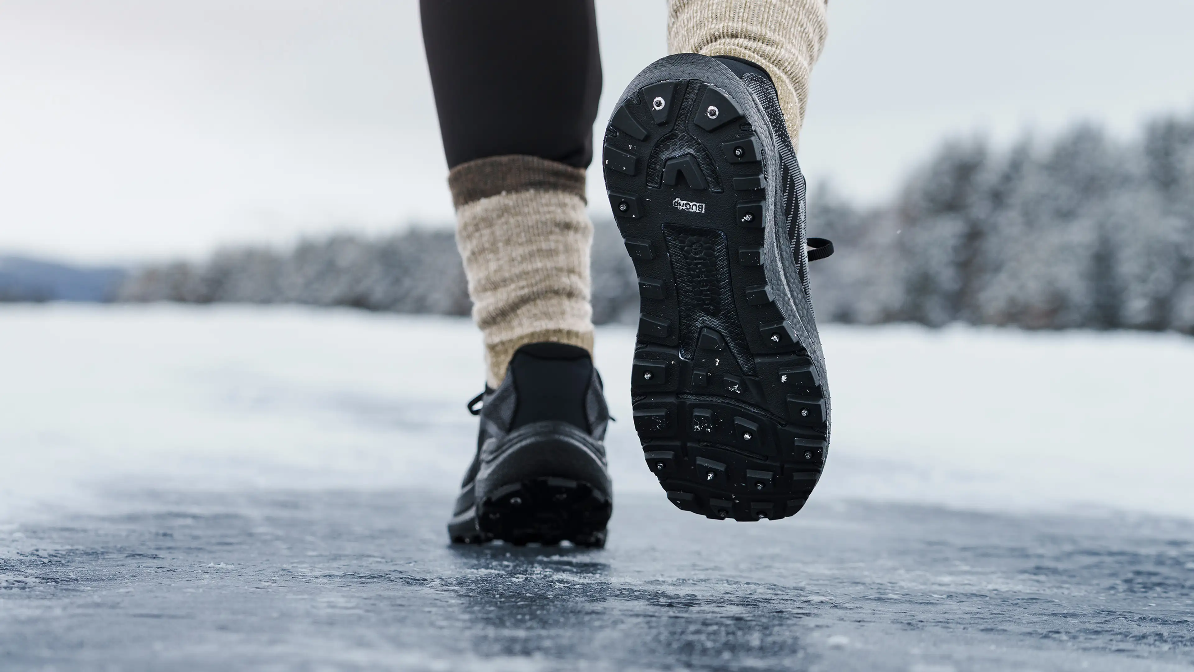 Close-up of a person wearing spiked black shoes and beige socks, walking on an icy surface. Snowy landscape in the background.