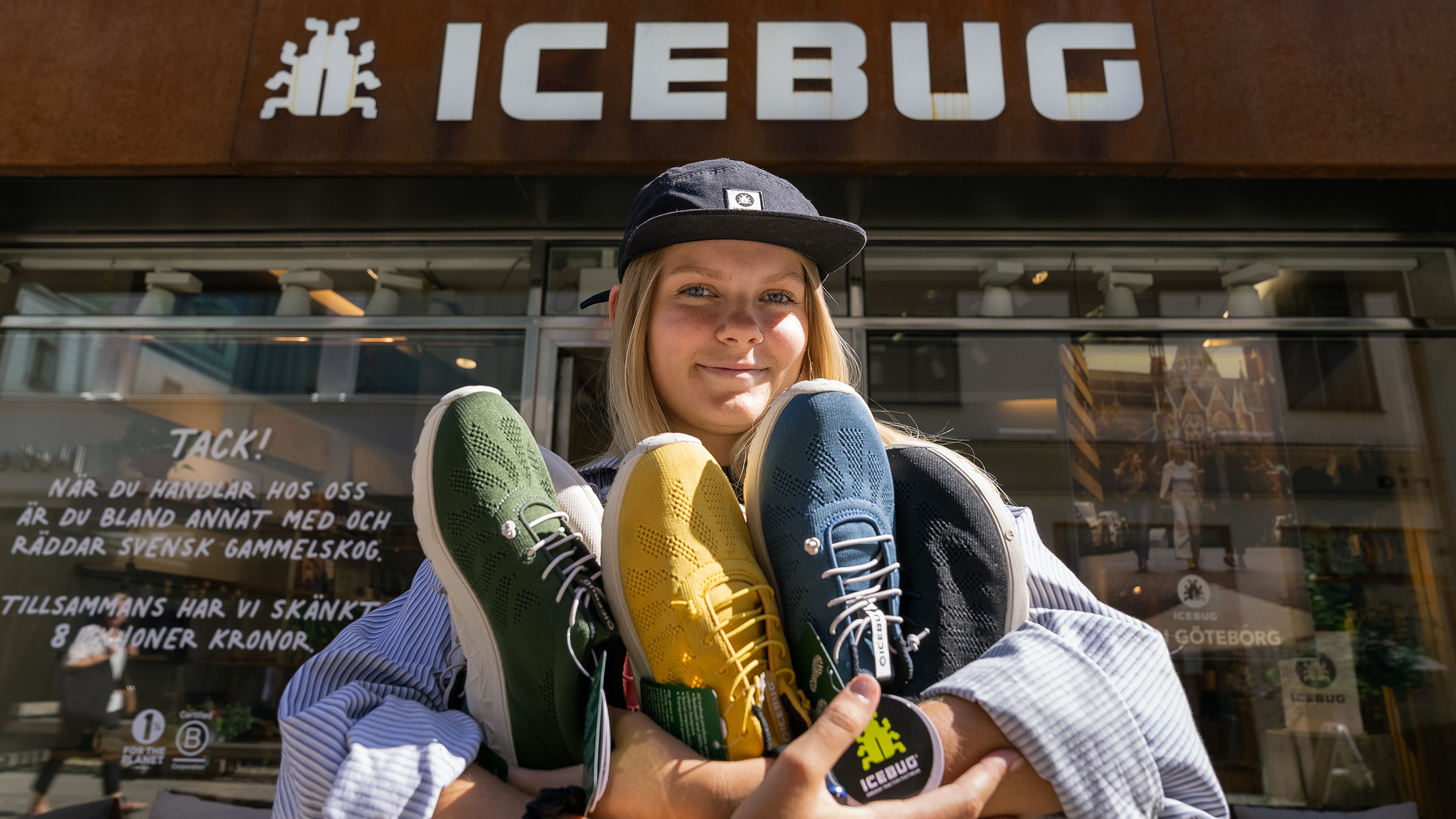 Person holding three colorful pairs of shoes in front of an Icebug store, wearing a cap and smiling.