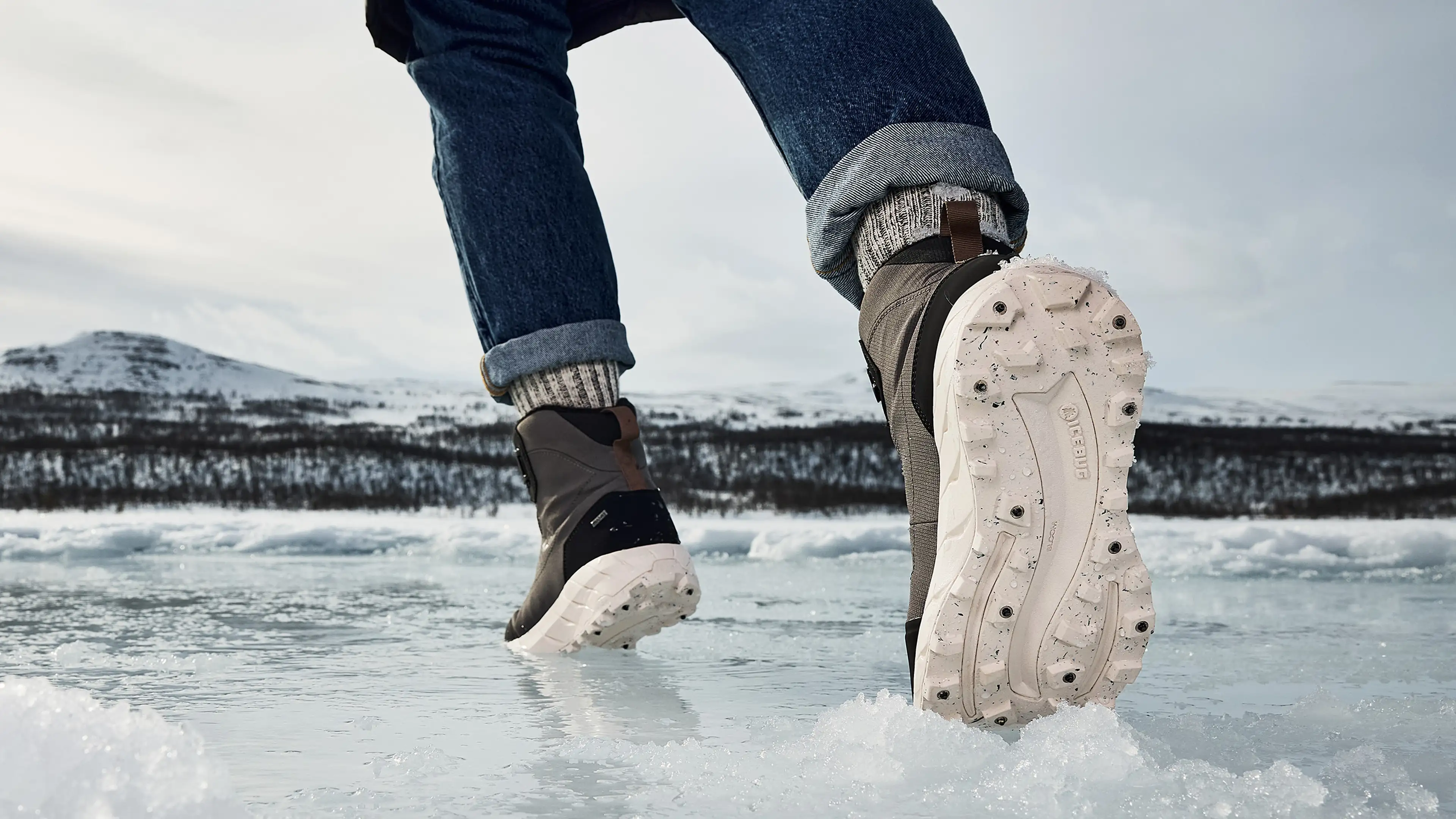 Person wearing Icebug winter boots and jeans walking on icy terrain with snow-covered mountains in the background.