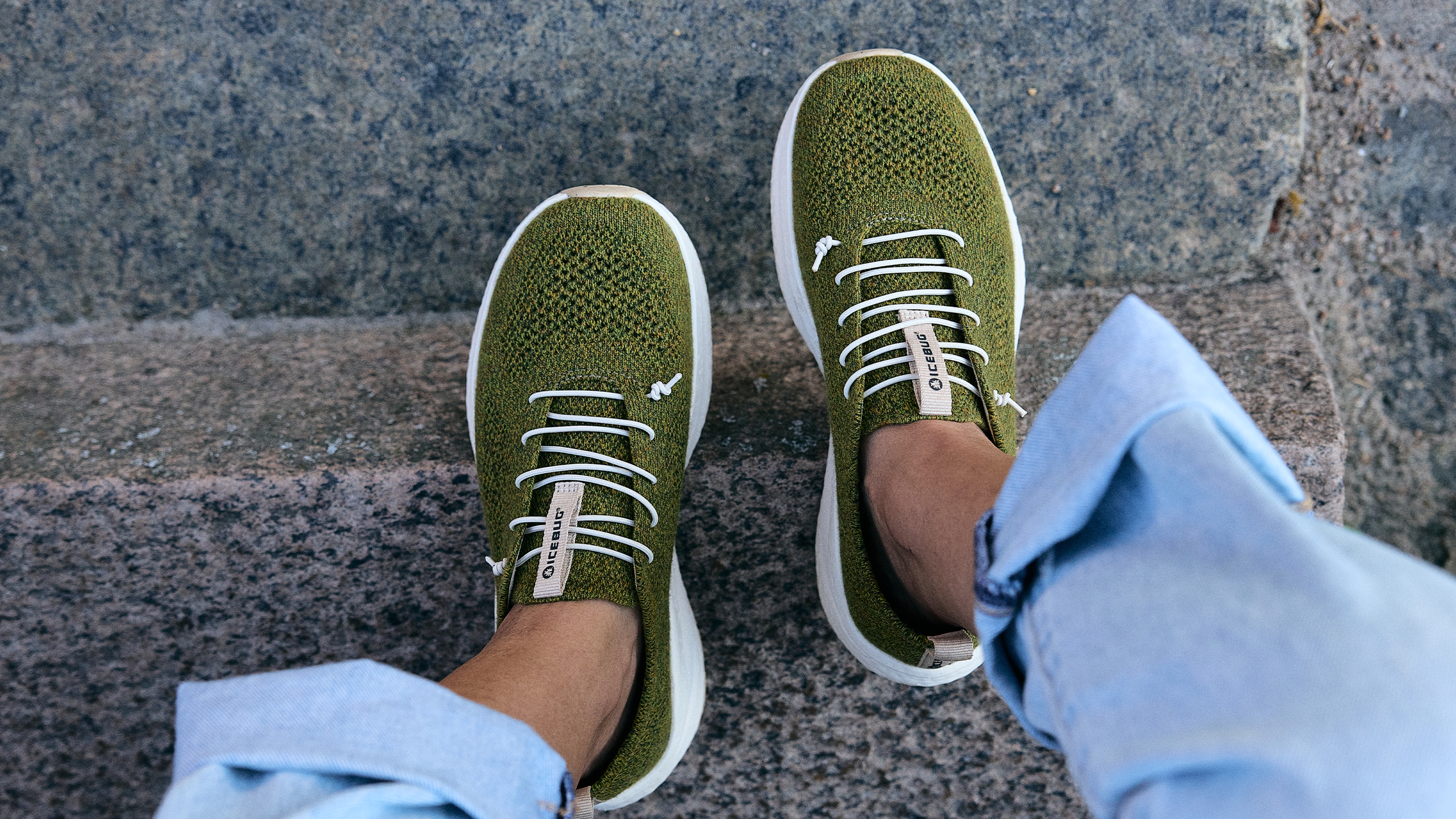 Person wearing green wool sneakers from Icebug with white laces, standing on stone steps, wearing light blue jeans.