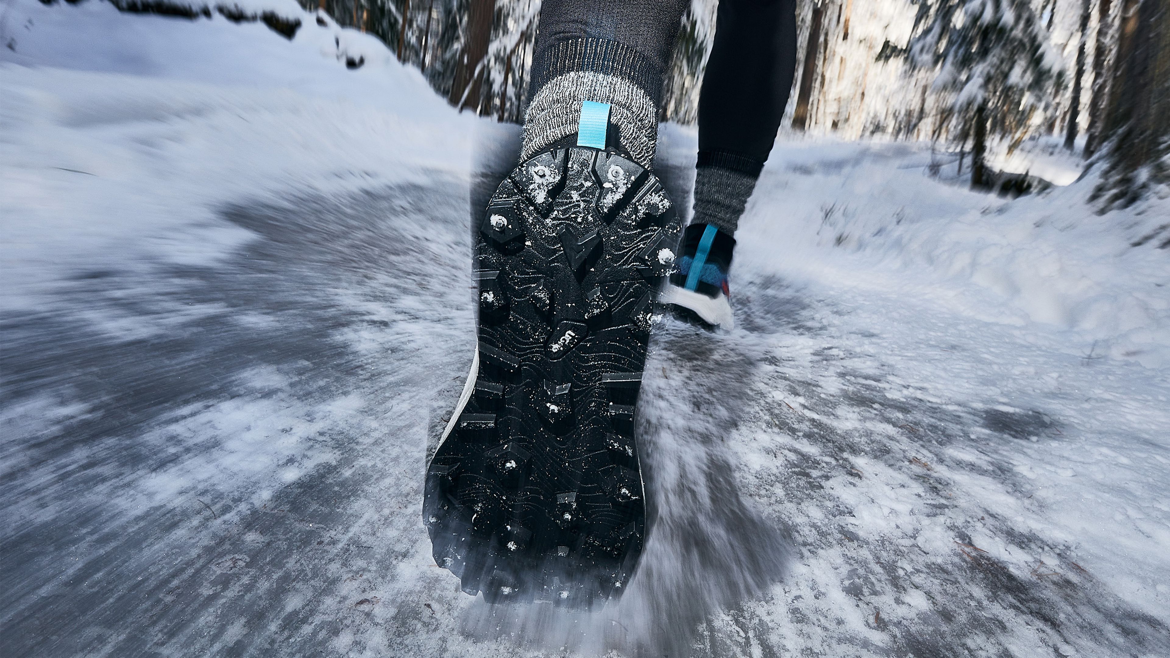 Close-up of a person running on an icy path, wearing Icebug shoes with spiked soles for grip, surrounded by snow-covered trees.